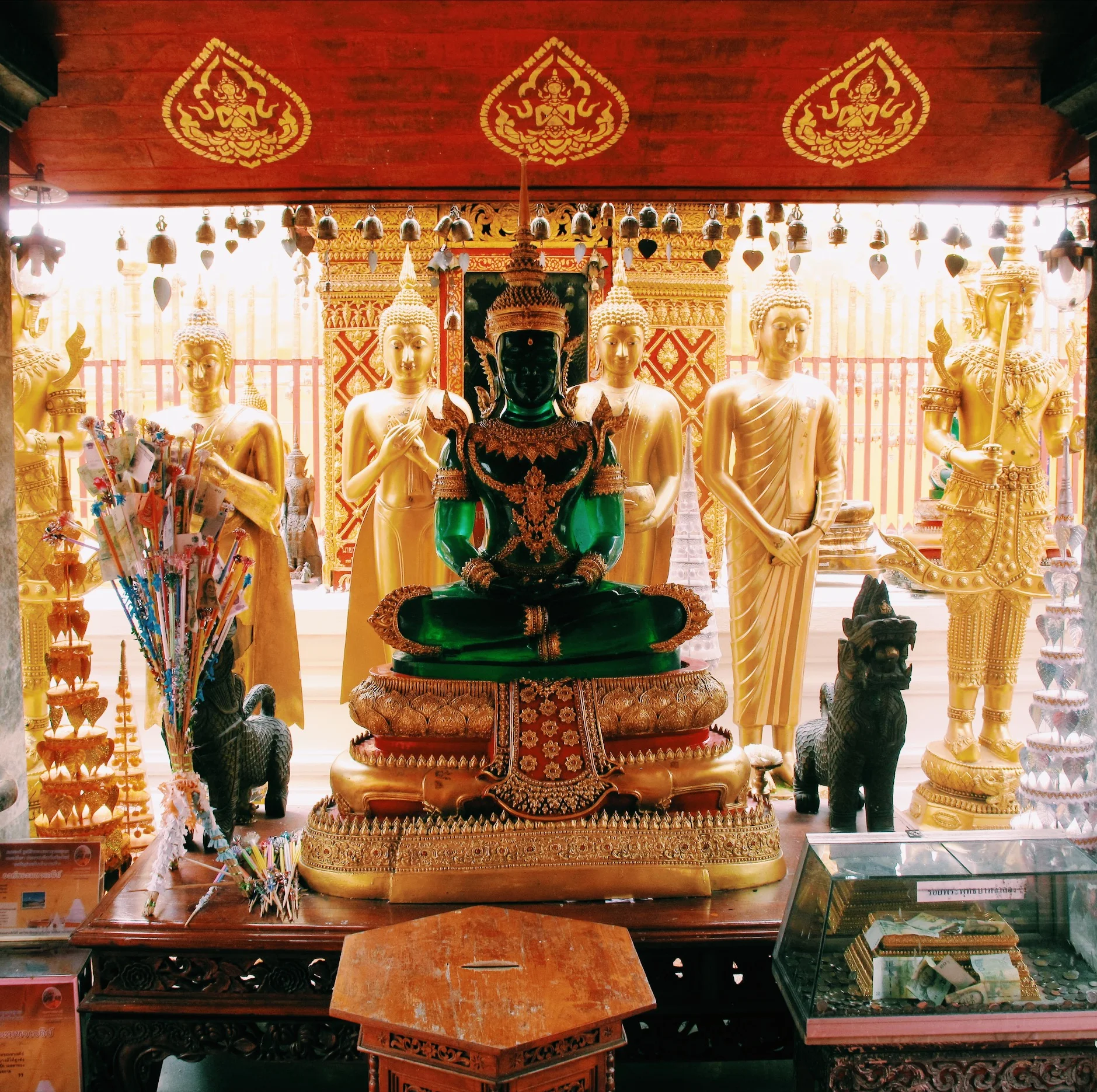 A temple interior with golden Buddha statues and a central green deity statue, intricately decorated and surrounded by offerings, in an ornate setting.