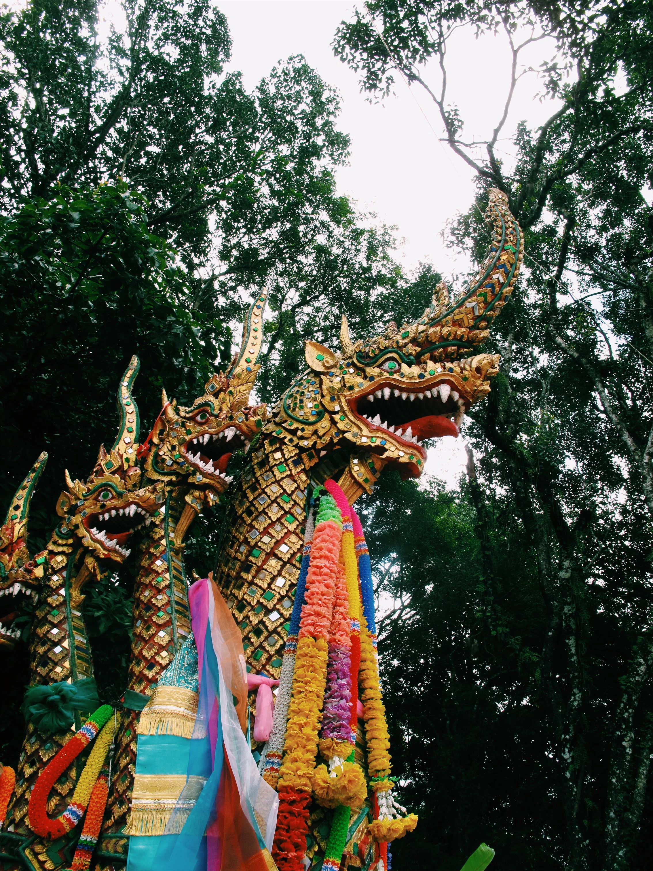Decorative Naga serpent statues adorned with colorful garlands in a lush forest setting.