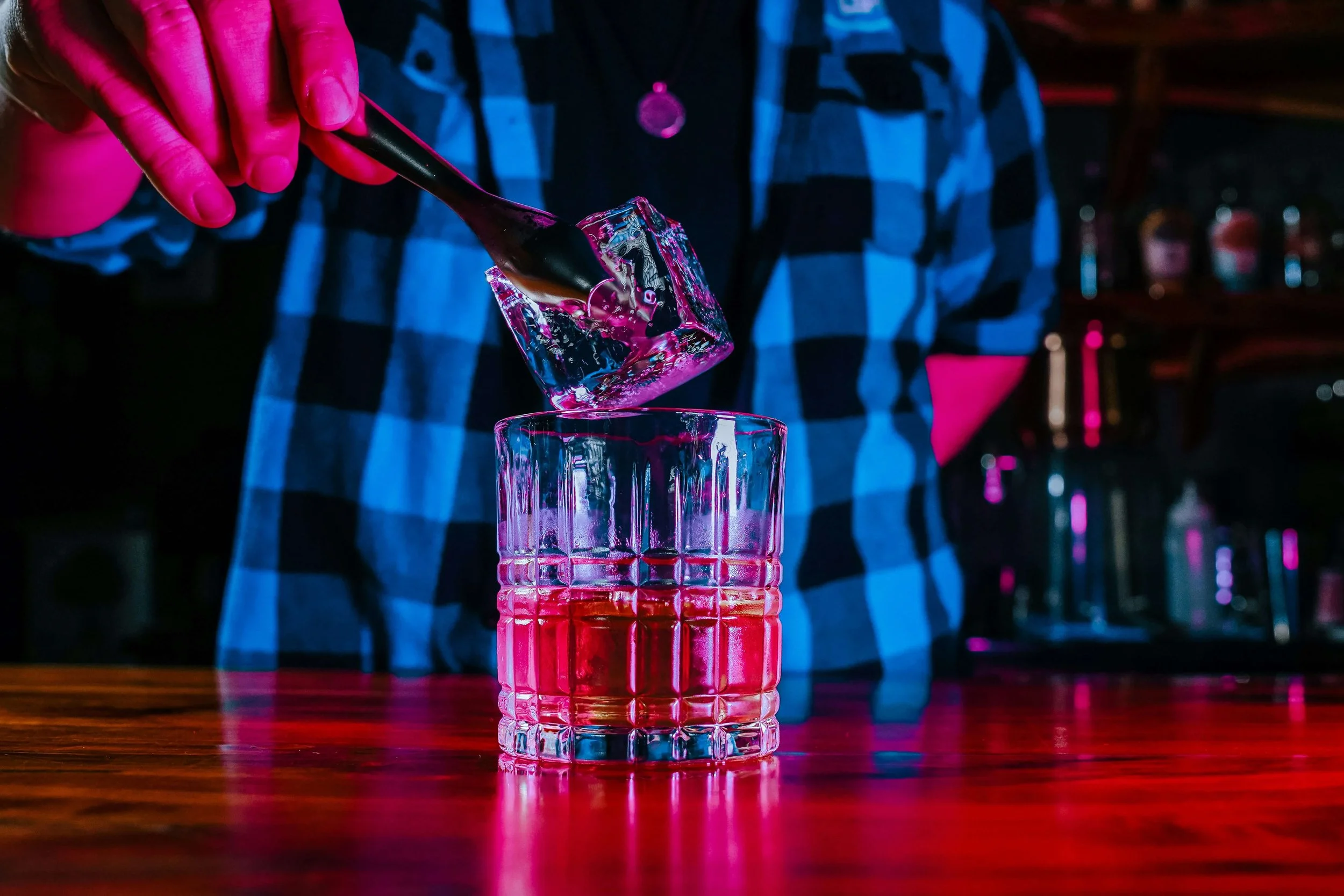 A person in a blue and black checkered shirt is pouring a clear liquid into a glass with a patterned surface, set on a wooden bar or table, in a dimly lit environment with colorful lighting.