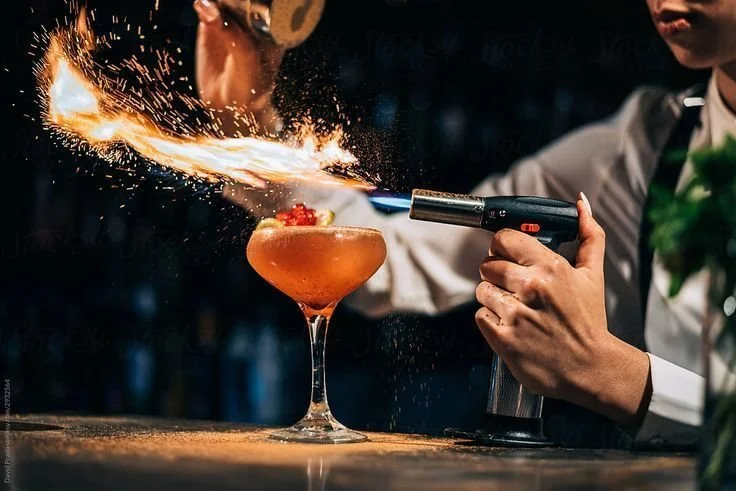 A bartender uses a torch to create a fiery display over a cocktail in a glass. The bartender is wearing a white shirt with a green tie, and the background is dark.