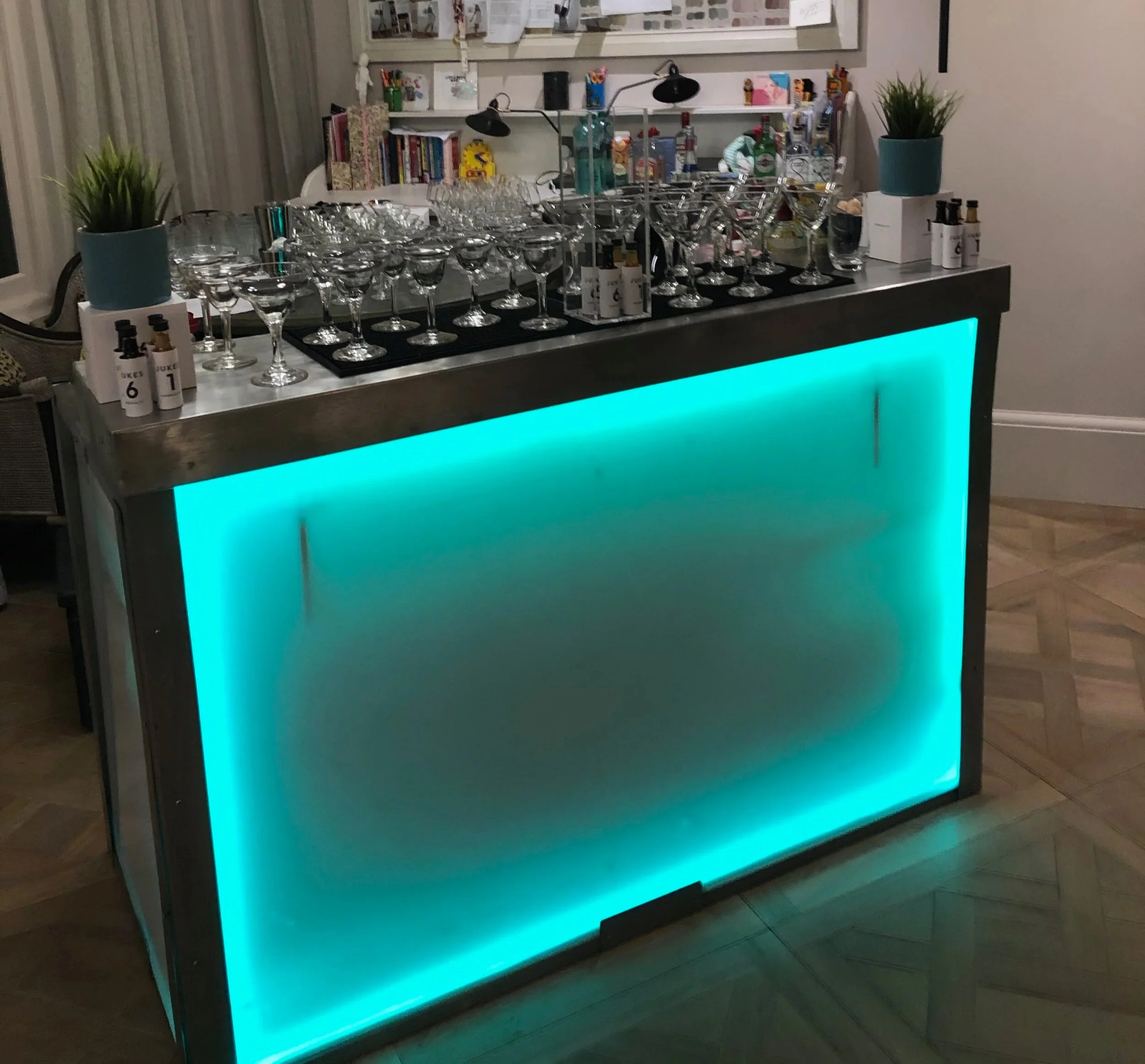 A bar counter with a turquoise illuminated front panel, topped with various glasses and bottles, and decorated with potted plants.