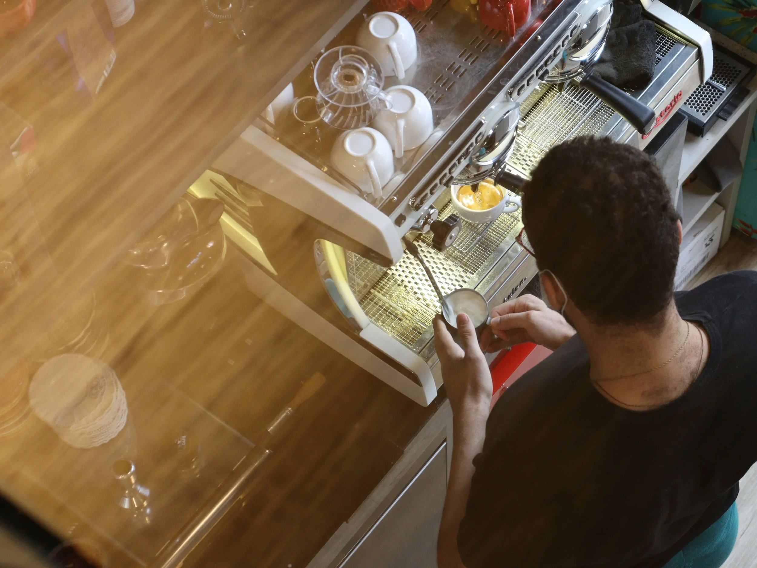 A person making espresso at a coffee shop counter, with equipment and cups on top of the machine, seen from above.