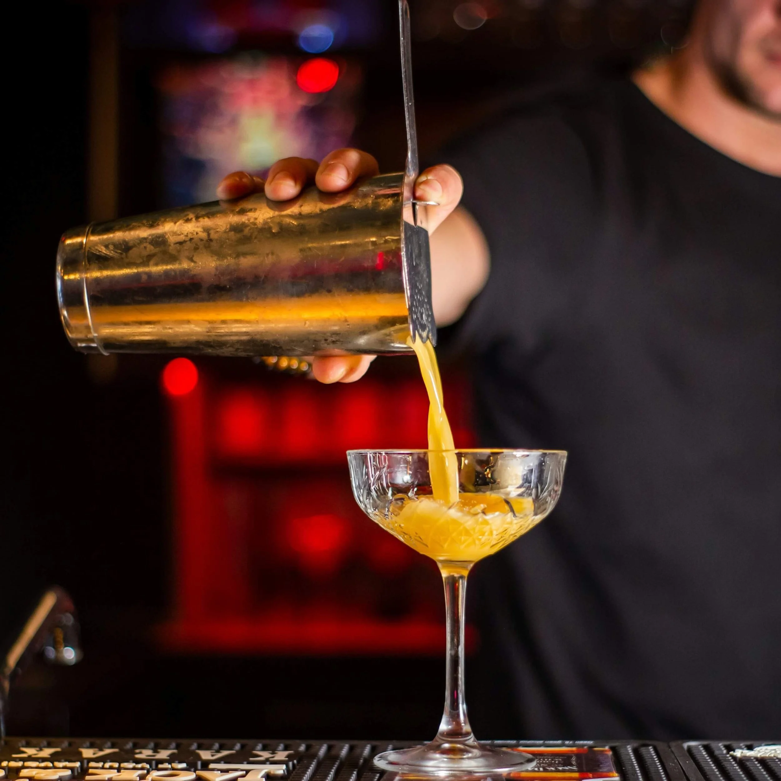 A bartender pours a yellow-orange cocktail from a shaker into a glass with a stem in a bar setting with colorful lights in the background.