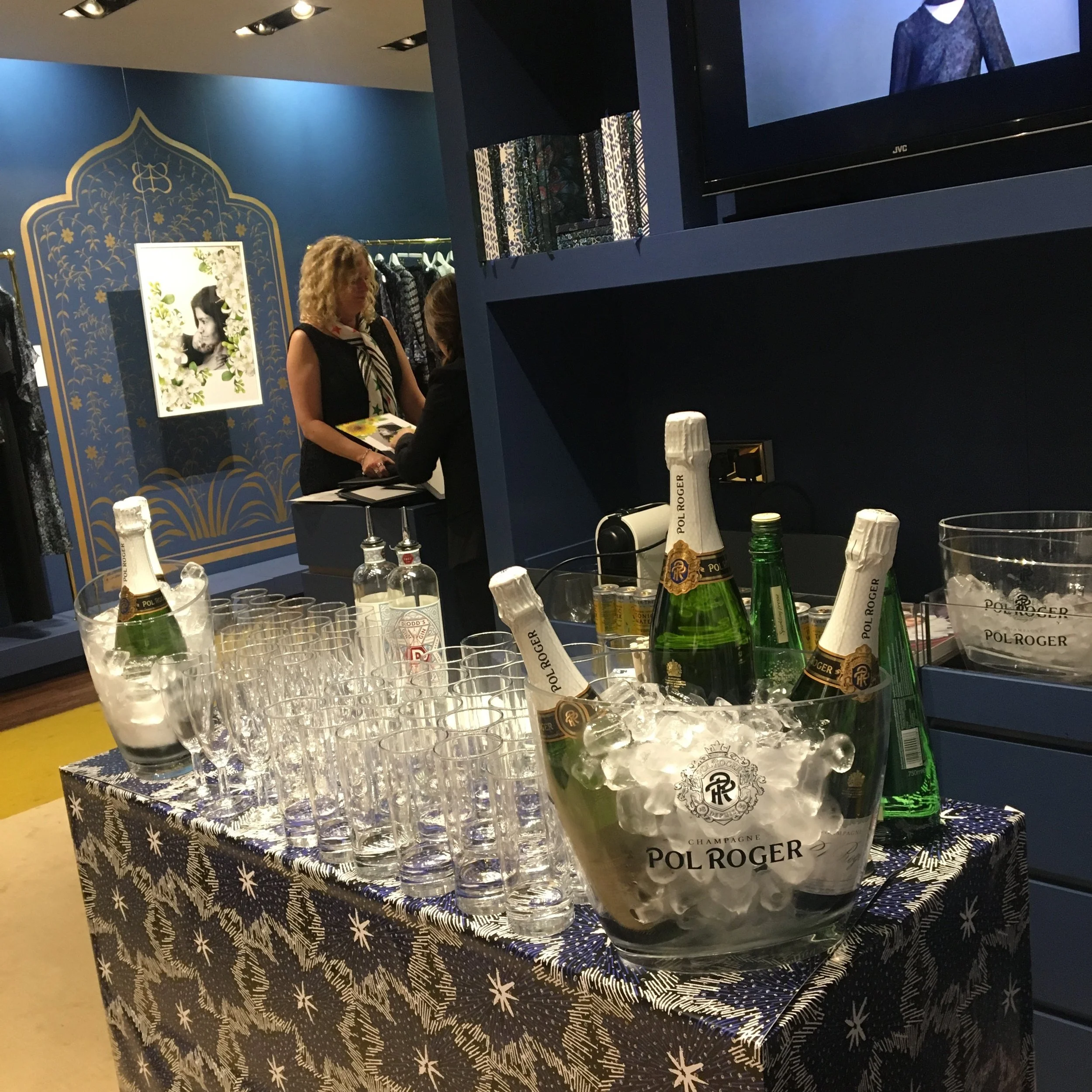 Table with bottles of champagne, glasses, and ice at a store event, with two women talking in the background.