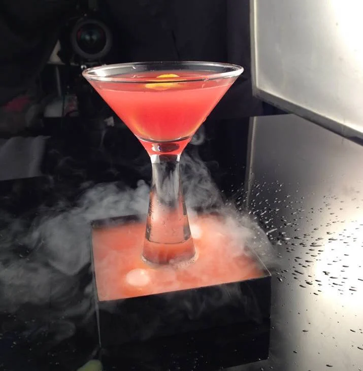 A pink cocktail in a martini glass on a black stand with dry ice and fog surrounding it.