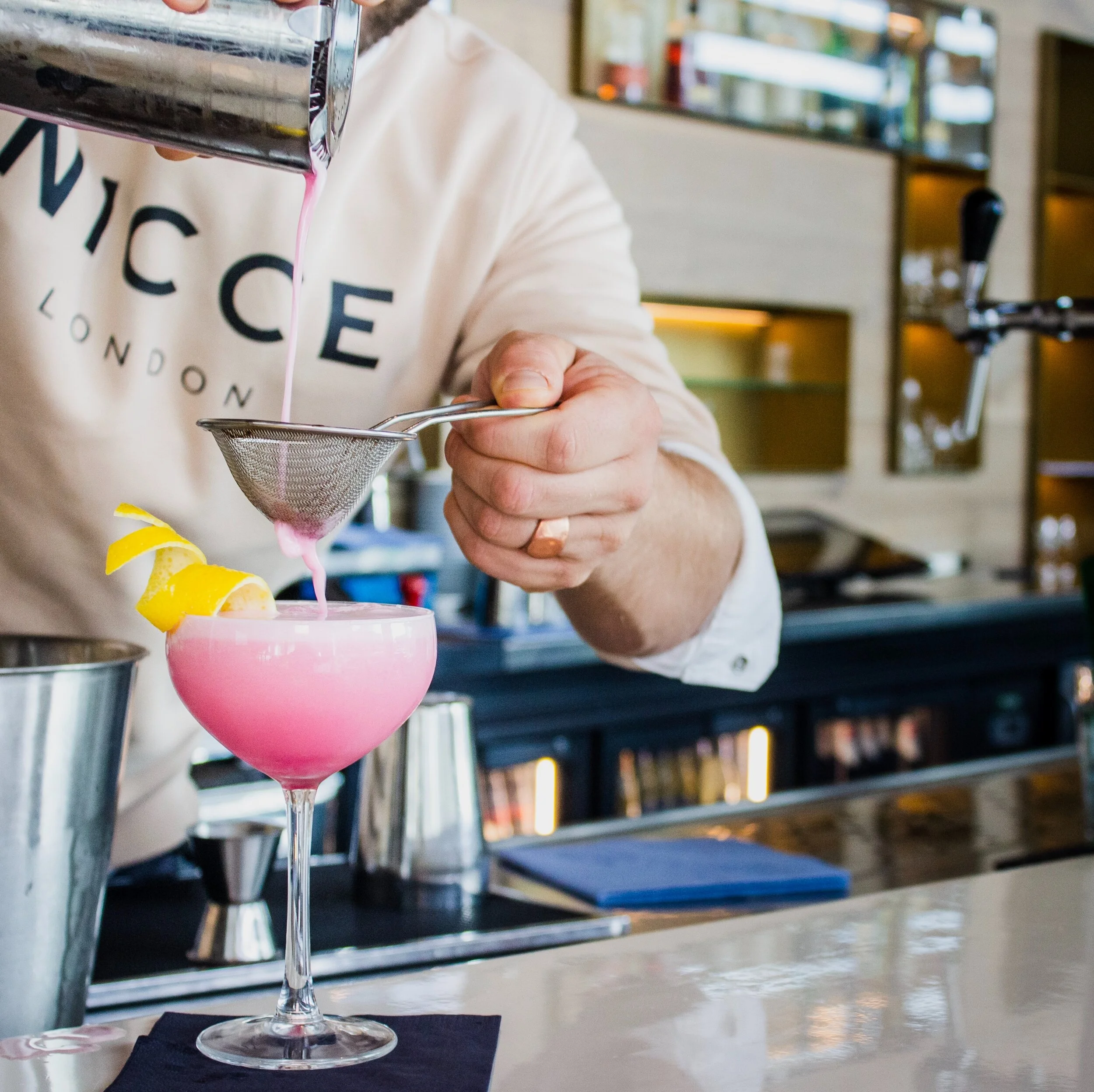 A bartender is straining a pink cocktail into a coupe glass garnished with lemon peel at a bar.