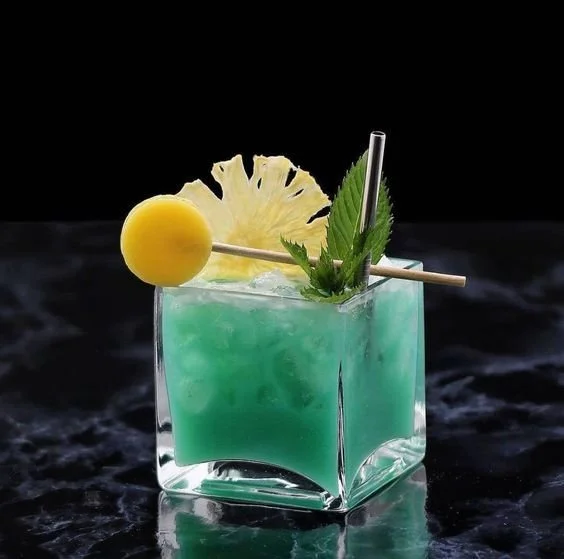 A vibrant blue-green cocktail in a clear square glass, garnished with pineapple slices, a lemon ball on a skewer, and a sprig of mint, with two straws.