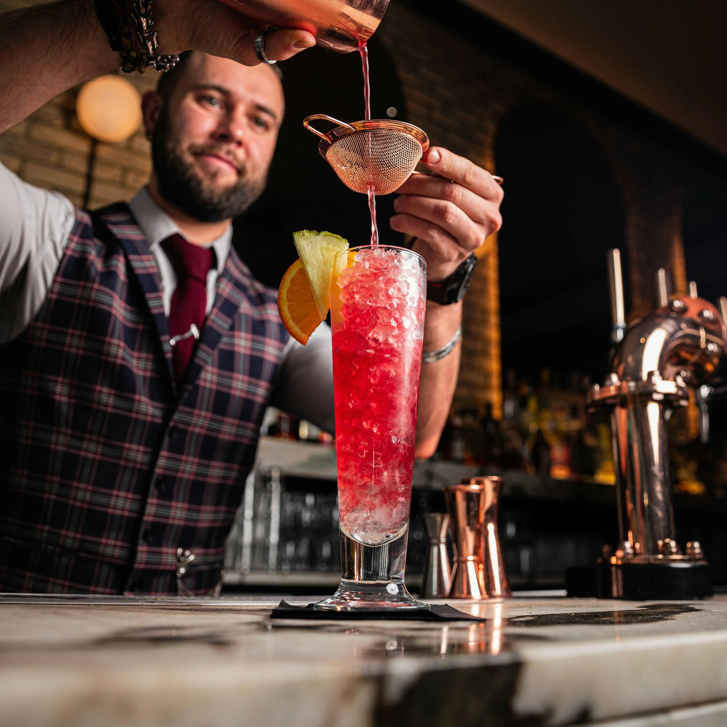 Bartender making a cocktail with crushed ice, garnished with a lemon and orange slice, at a bar with brick wall and copper accents.