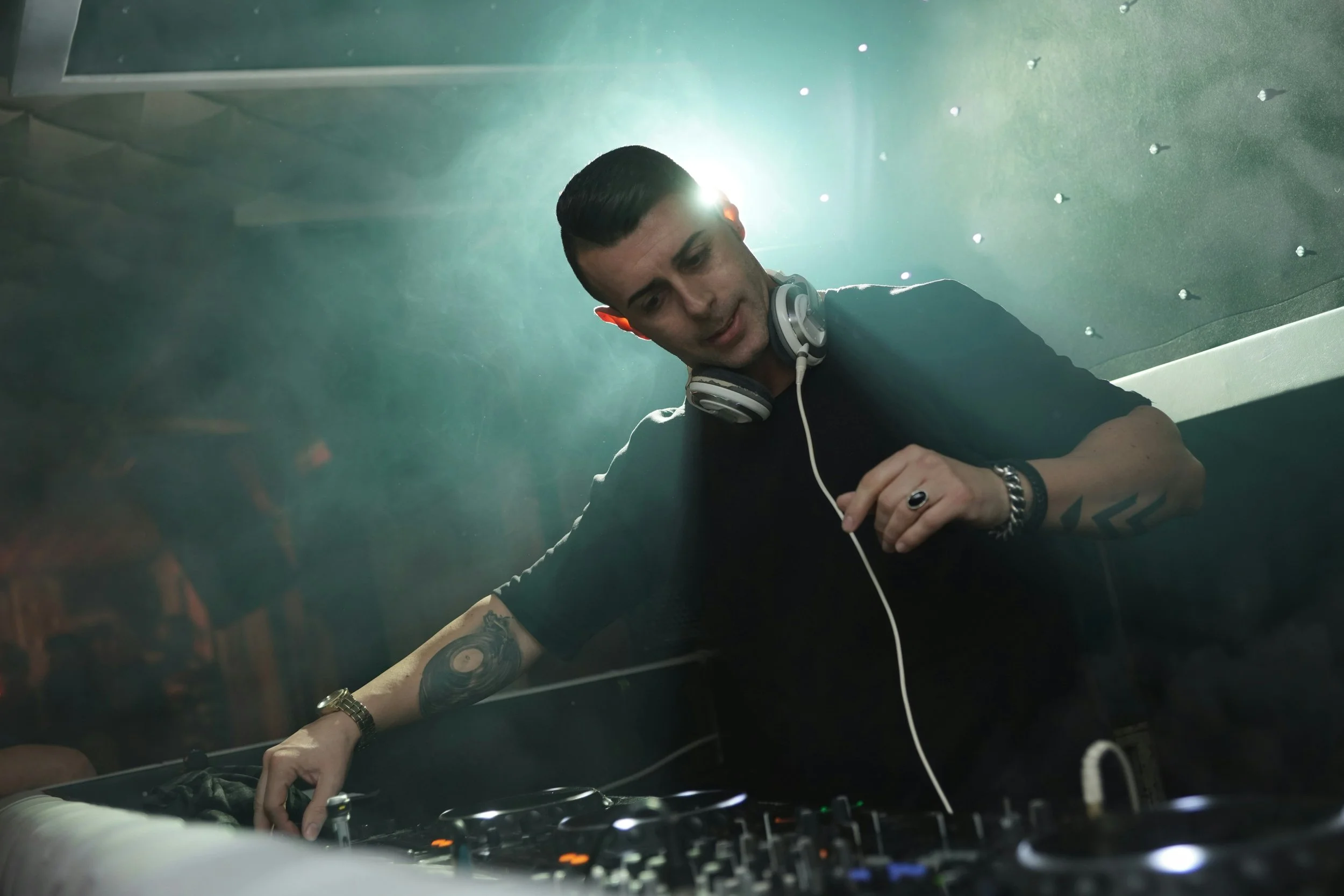 A male DJ wearing headphones is mixing music on a DJ console in a club or concert setting, with green lighting and a foggy atmosphere.