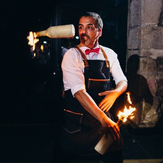 A man in a white shirt, red bow tie, and apron is balancing a flaming baton on his shoulder and holding another lit baton in his hand in front of a dark background.