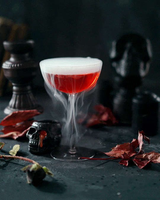 A cocktail glass with red drink and dry ice fog on a dark surface, surrounded by Halloween-themed decorations like skulls and dried leaves.