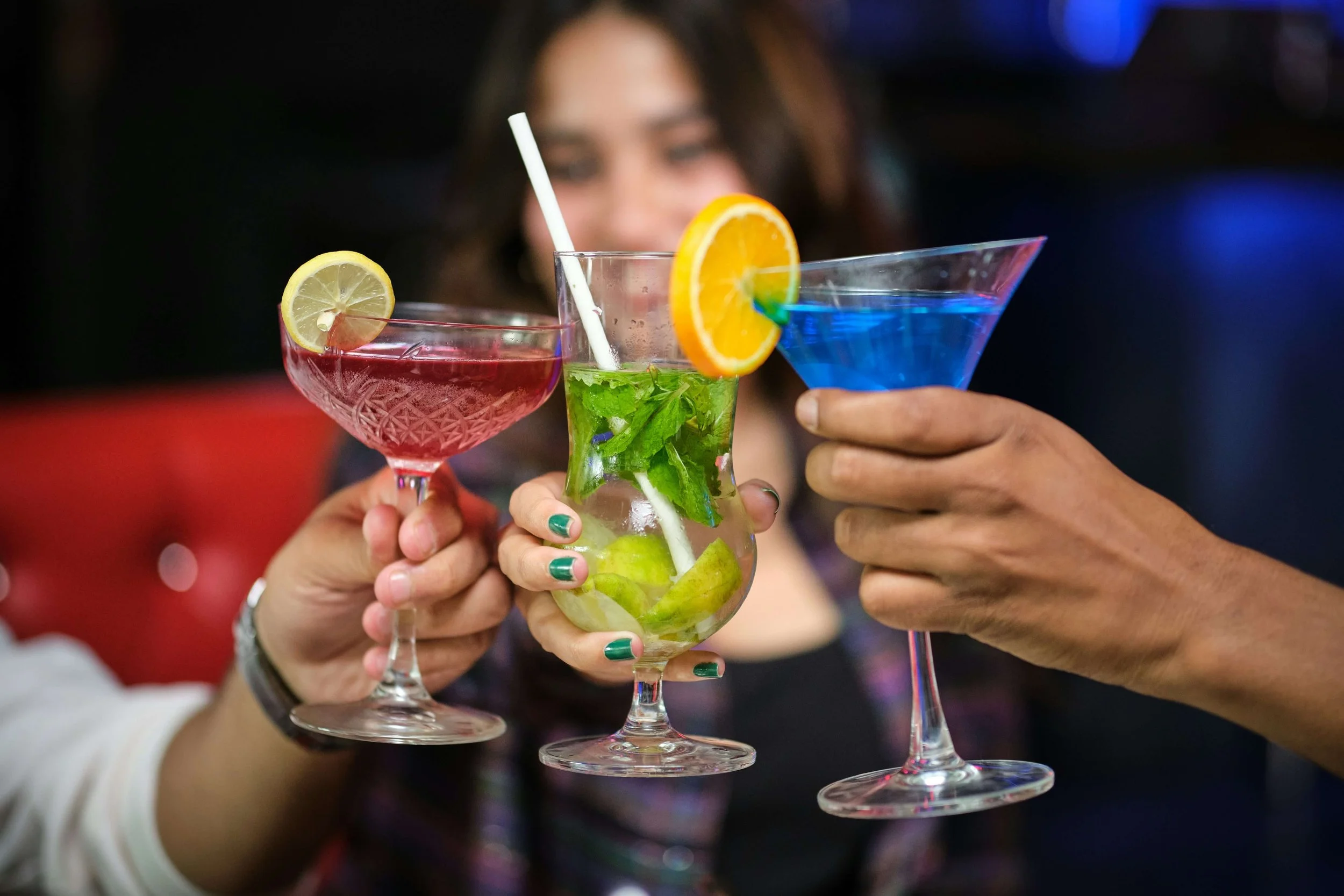 Three people holding colorful cocktails in a toast, with a woman in the background. The drinks include a pink cocktail with a lemon slice, a clear mojito with mint and lime, and a blue cocktail in a martini glass.