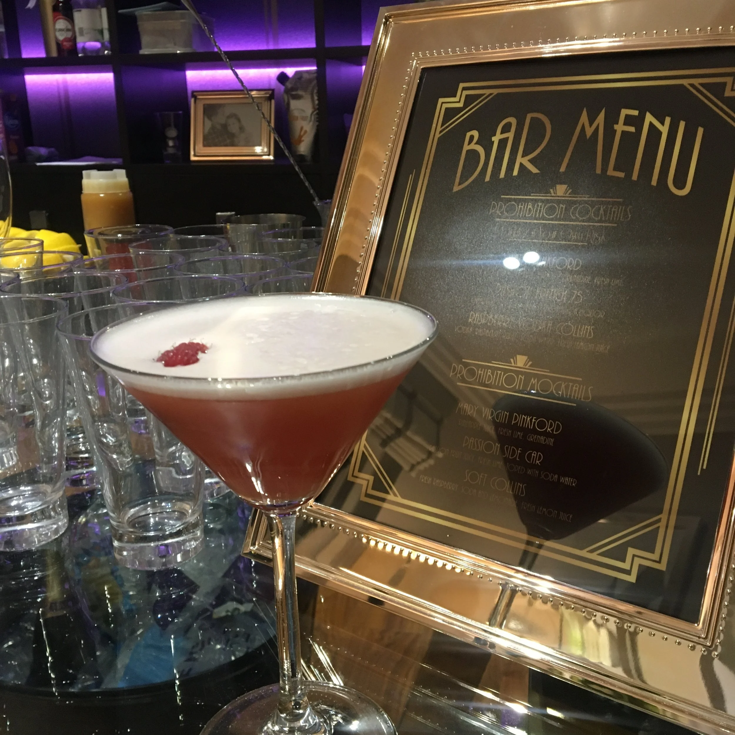 A martini glass with a pink cocktail and a cherry garnish on top, placed on a bar counter next to a framed bar menu, with glasses of water and bar supplies in the background.