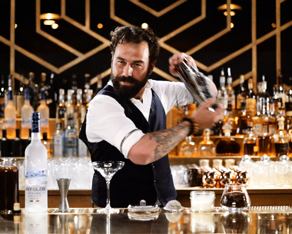 Bartender with a beard and tattoos pouring a drink at a bar with whiskey bottles in the background.