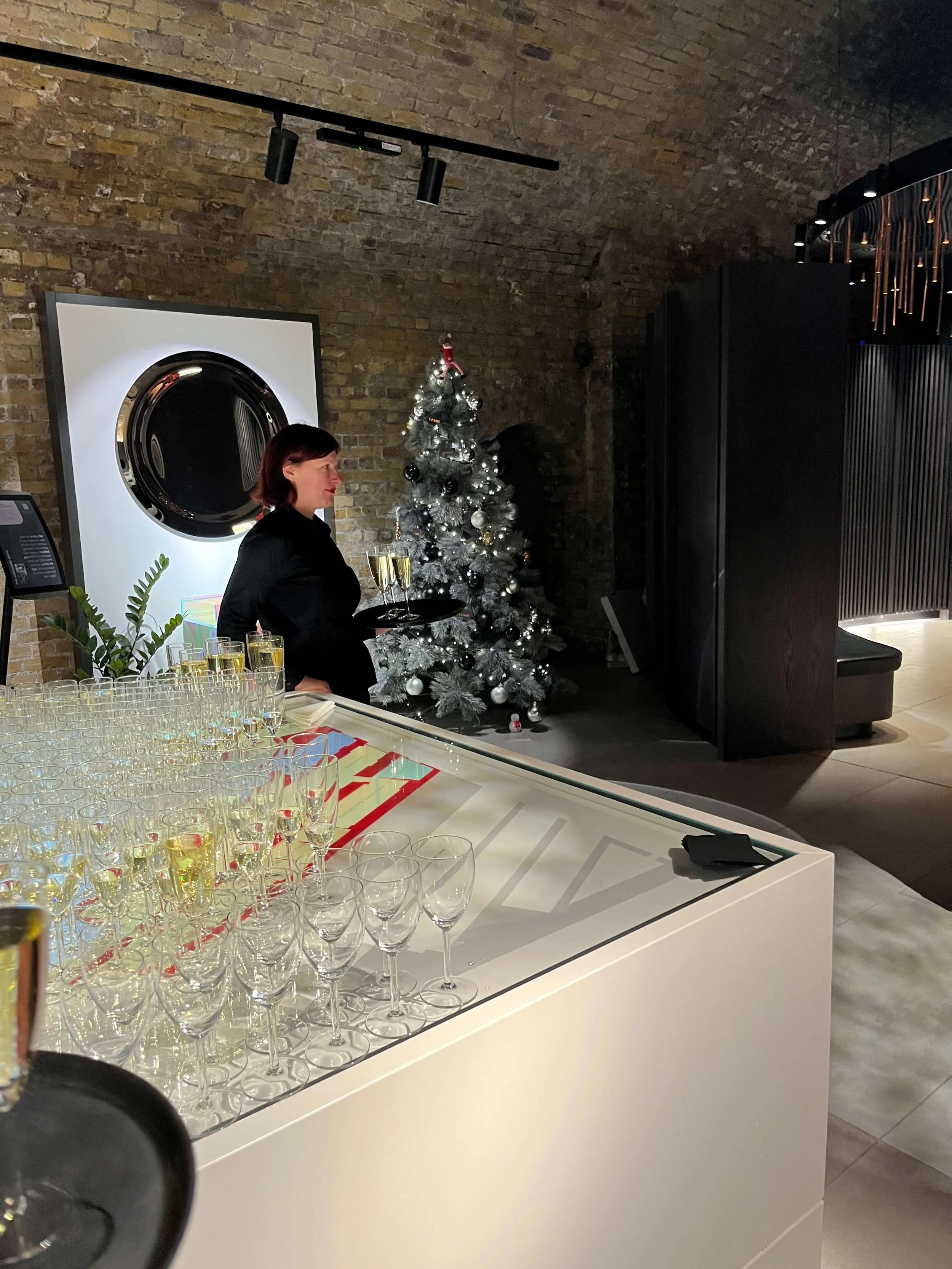 A woman in black holding a tray of champagne glasses at a bar with a decorated Christmas tree in the background.