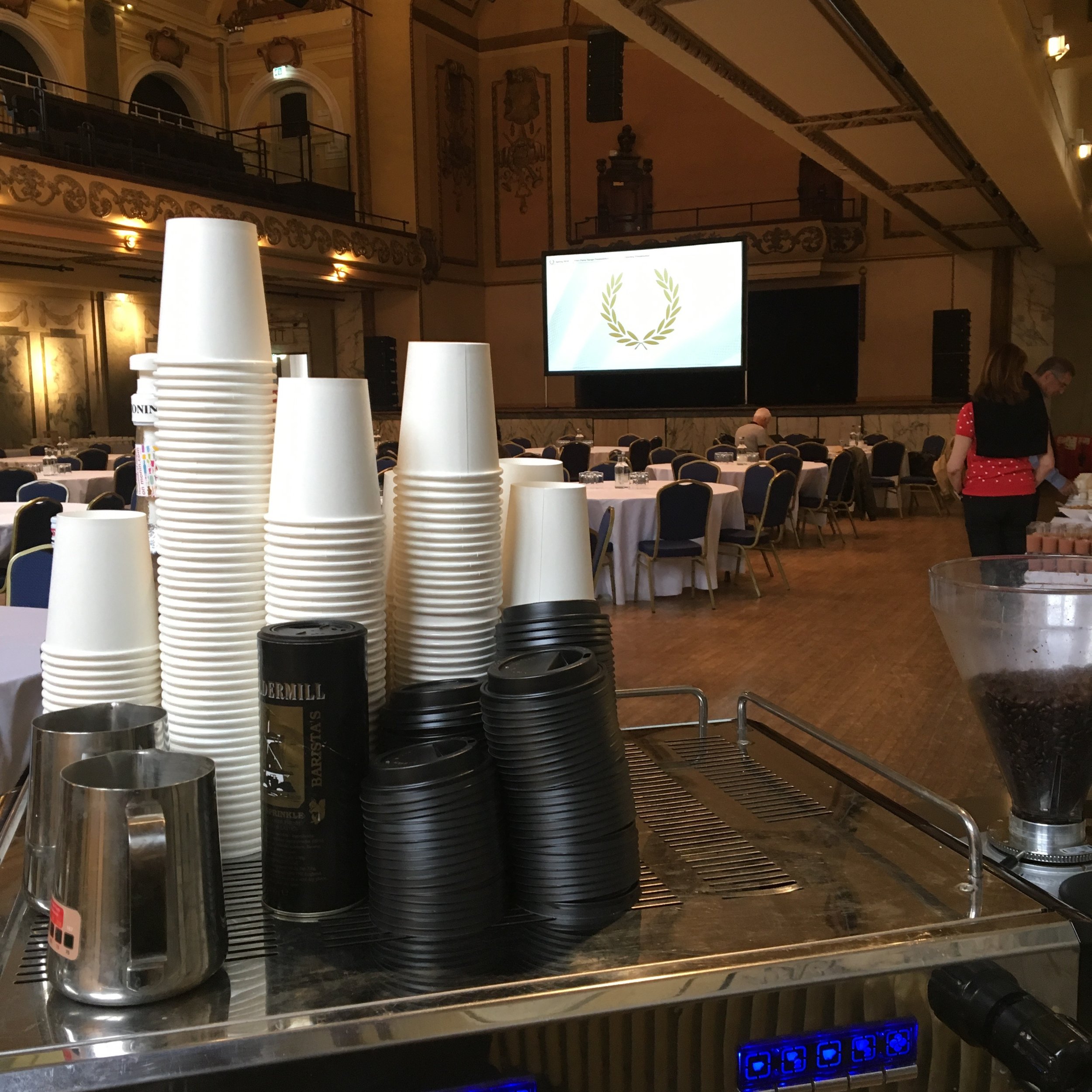 Stacked disposable cups, coffee stirrers, and containers on a table in a large banquet hall with round tables and a large screen displaying a laurel wreath emblem.