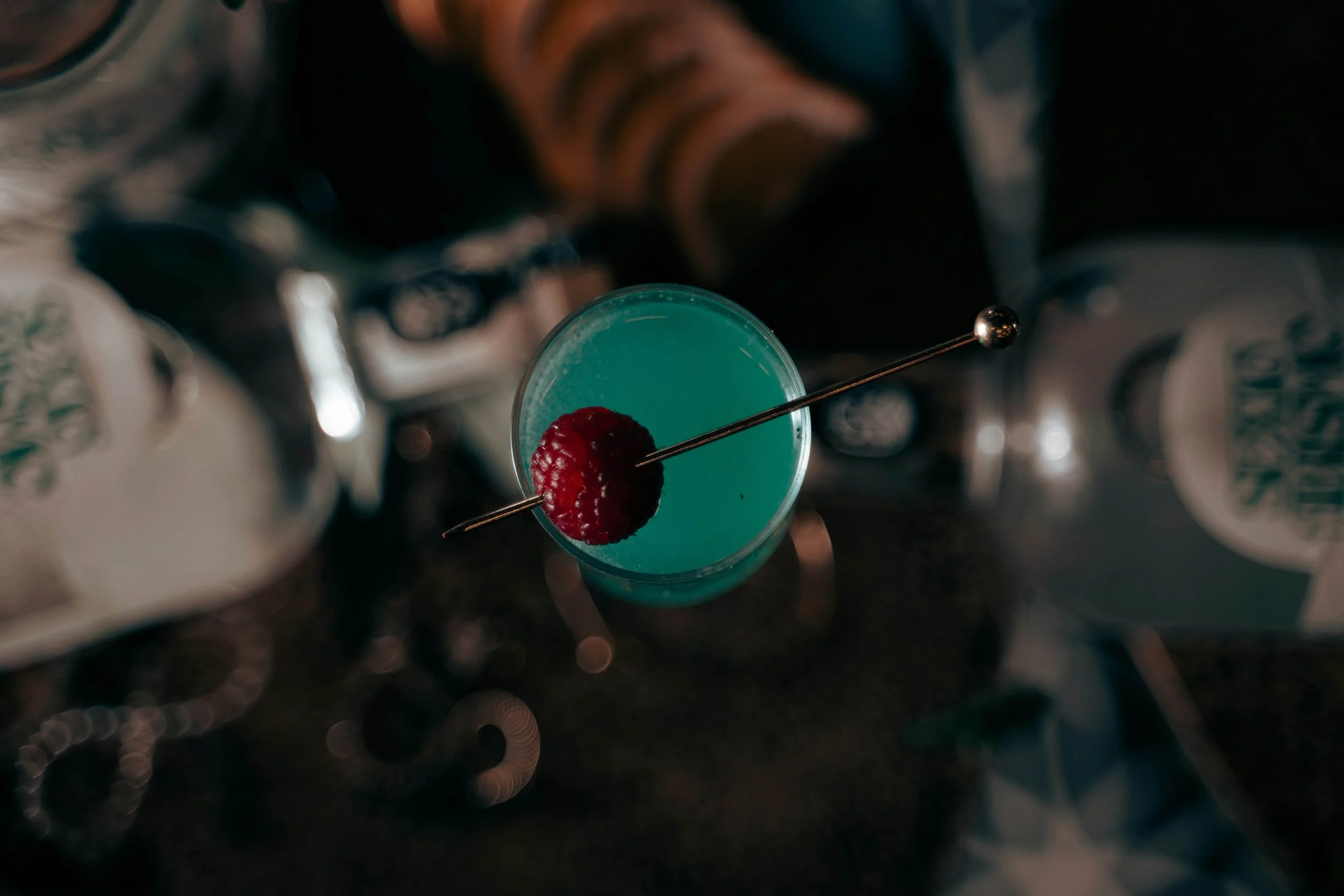 Overhead view of a blue cocktail garnished with a raspberry, with various bottles in the background.