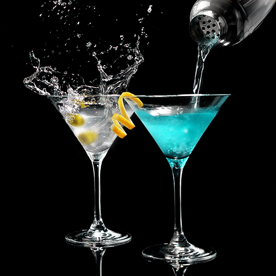 Two cocktails in martini glasses, one with a lemon twist, the other being mixed with blue liquid from a shaker.