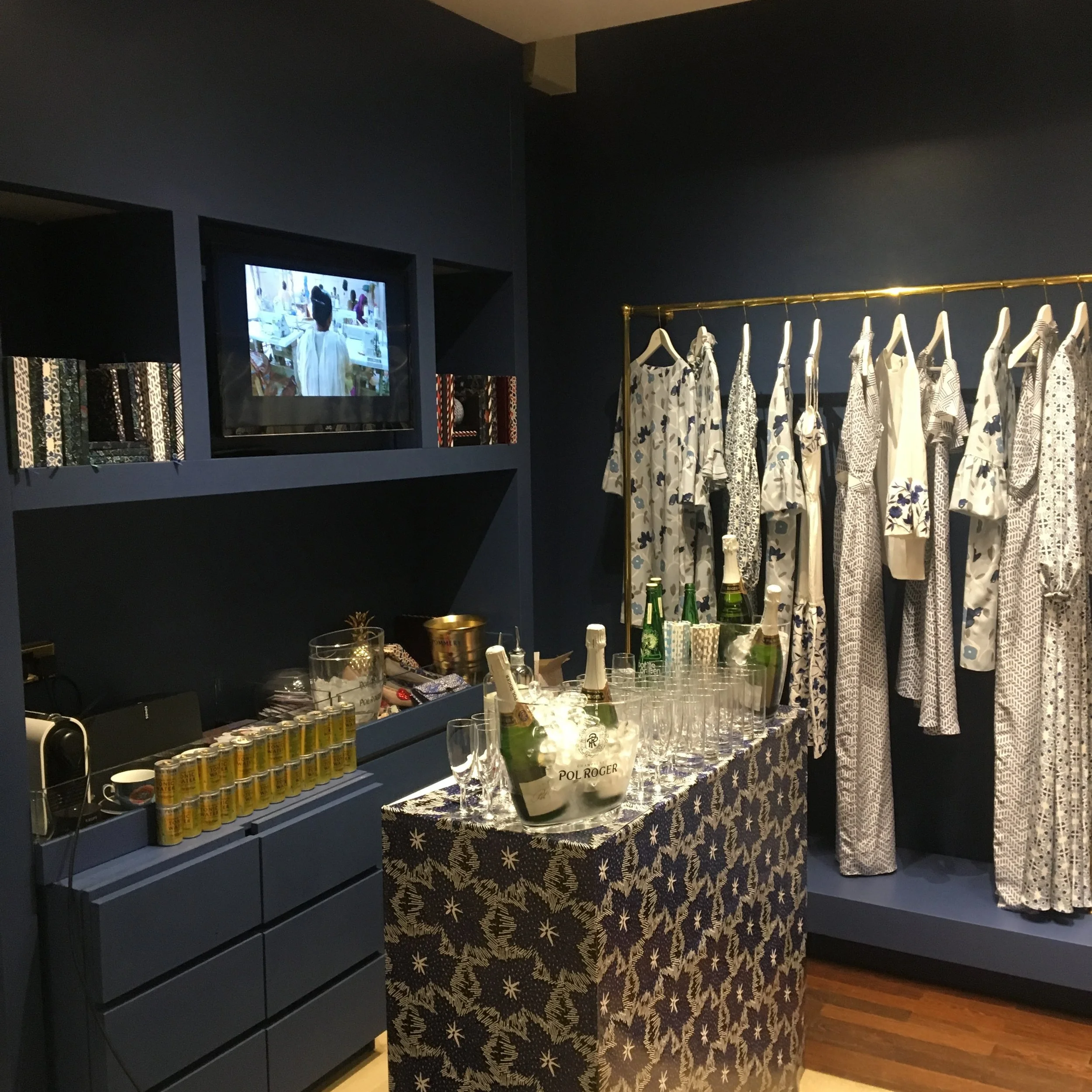 Display of blue and white patterned dresses and tops on a clothing rack, with bottles of champagne, glasses, and a tablecloth-covered table in a boutique.