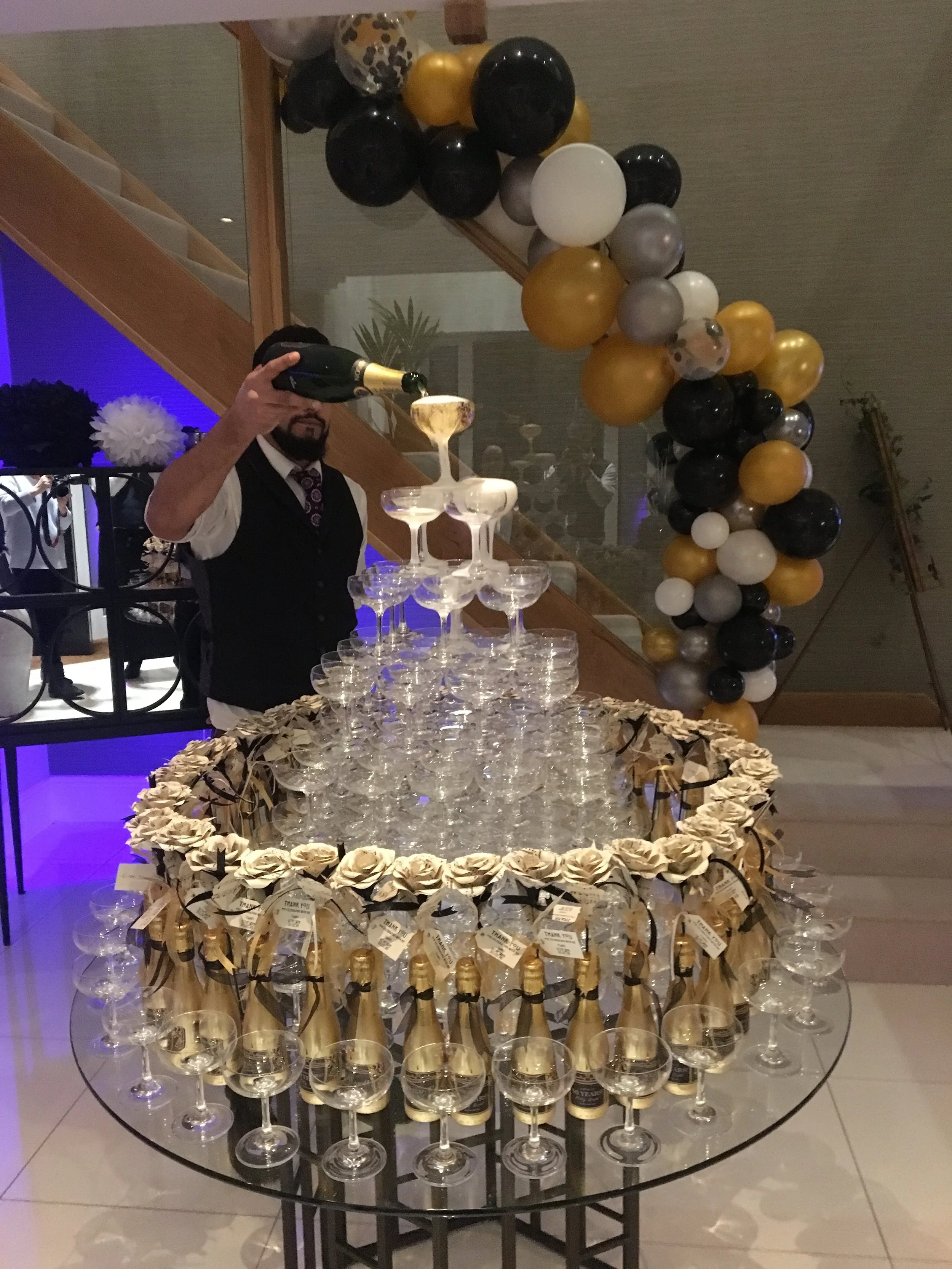 A man in a vest and tie pours champagne into a pyramid of glasses at a celebration with black, gold, white, and gray balloons forming an arch in the background.