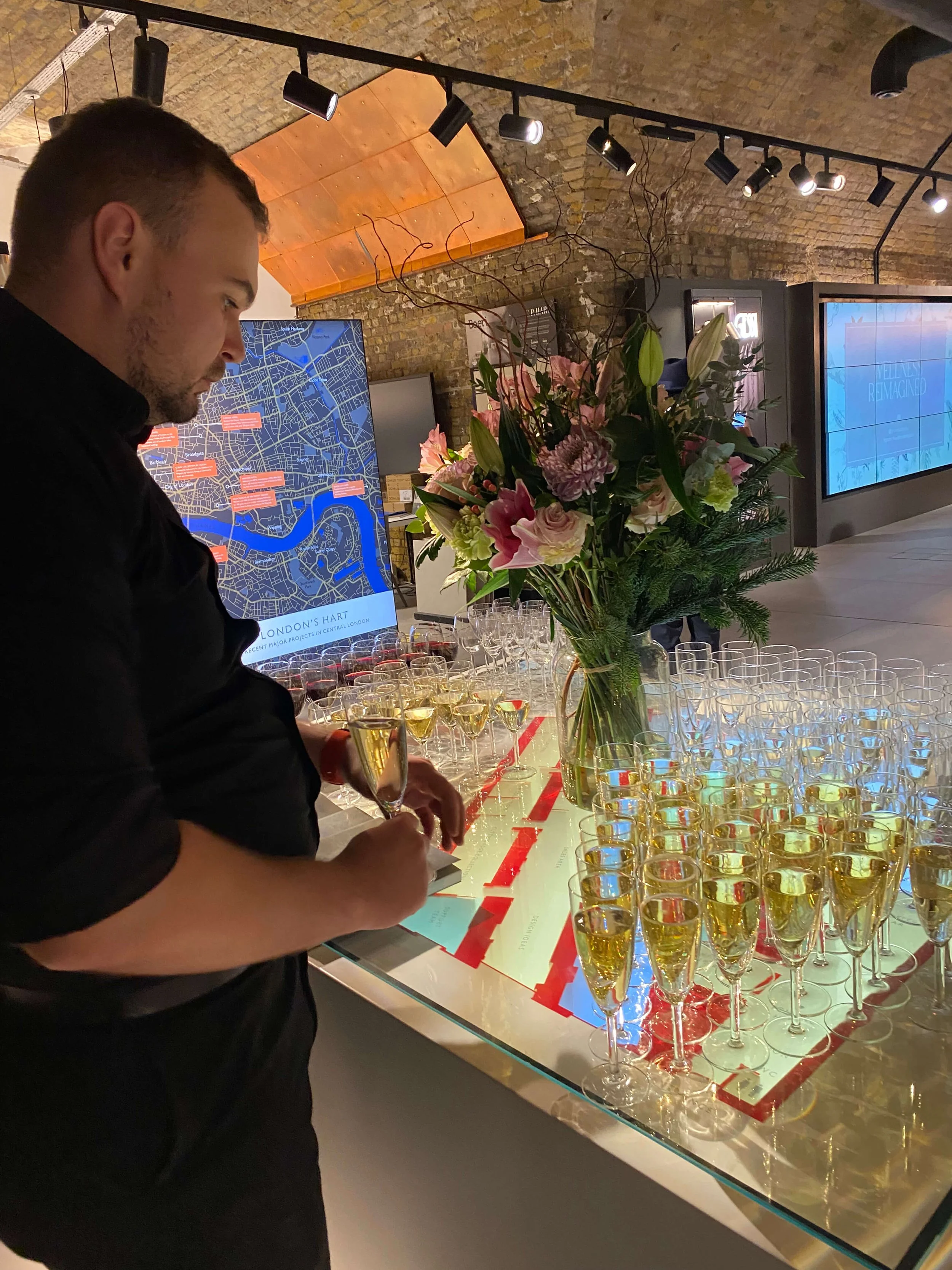 A man in a black shirt pouring champagne into glasses on a table decorated with pink and white flowers in a modern indoor setting with brick walls and digital displays.
