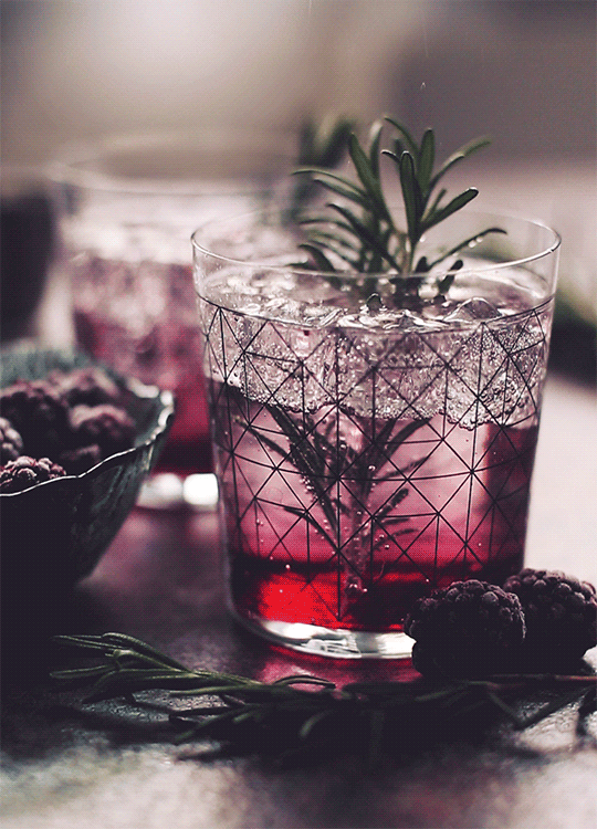 A glass of fruit-infused water with ice, garnished with a sprig of rosemary and surrounded by blackberries and additional rosemary sprigs.