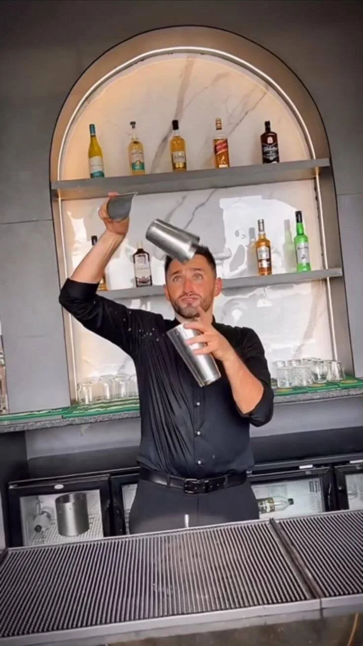 A bartender tossing a cocktail shaker in the air behind a bar counter with liquor bottles on shelves in the background.