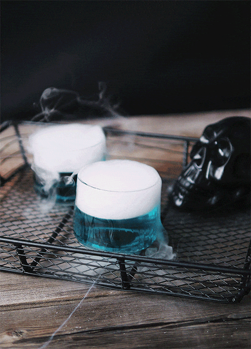 Two glass cups with dry ice creating fog inside, placed on a metal tray with a skull mask nearby.