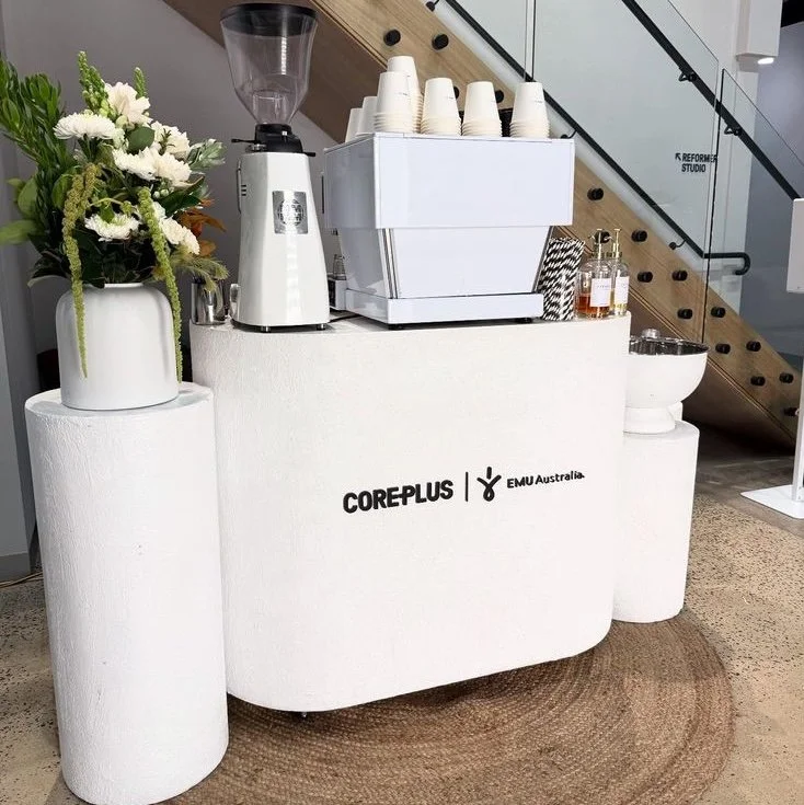 Coffee station with a coffee grinder, cups, and syrup bottles, decorated with a plant in a vase, on a white table with the words 'COREPLUS' and 'EMU Australia' printed on it, in a modern, minimalistic setting.