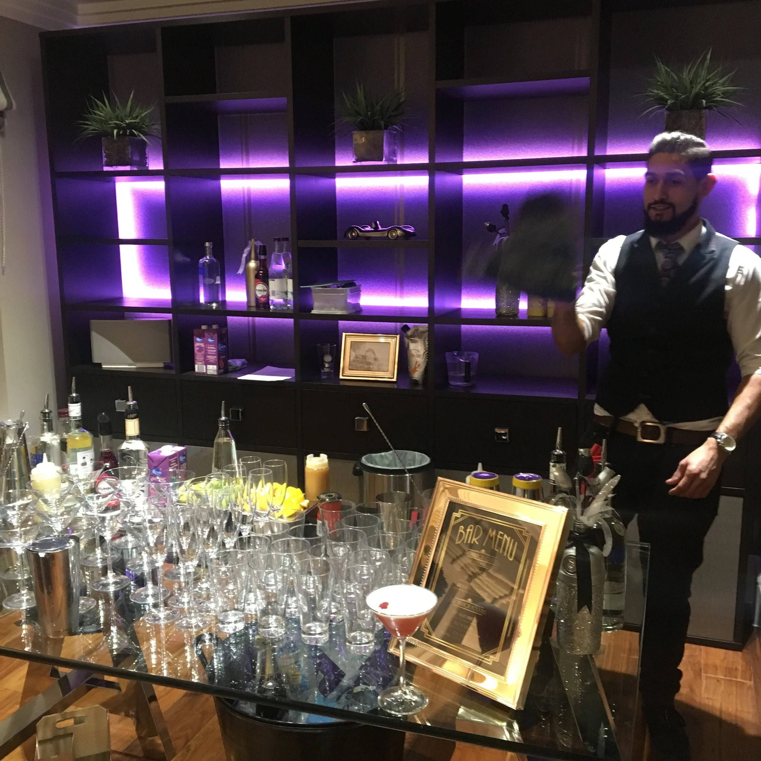 Bar area with bartender preparing drinks, glasses and cocktail ingredients on the table, illuminated wall shelves with bottles, plants, and decorative items.