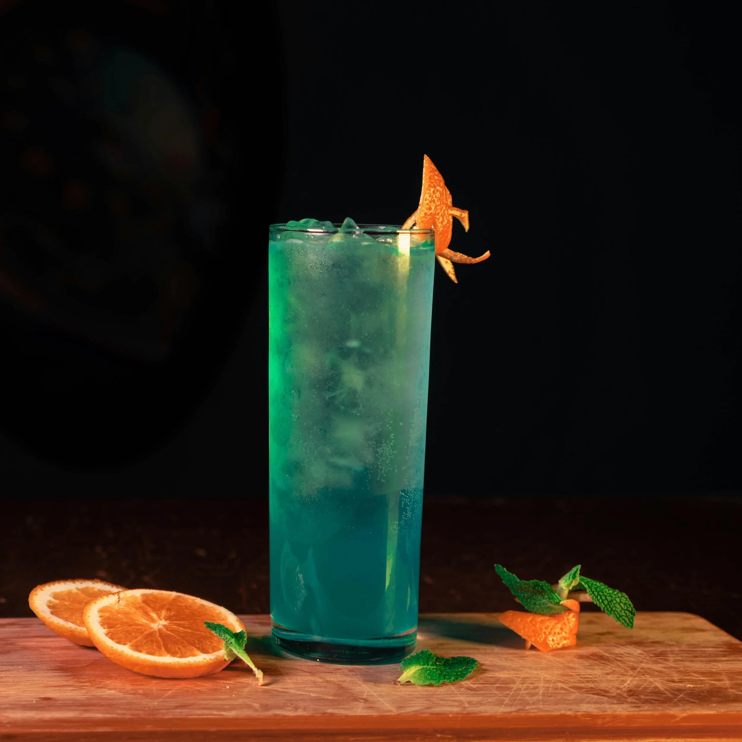 A tall glass of blue-green cocktail with ice, garnished with a twisted orange peel, on a wooden board with orange slices and mint leaves.