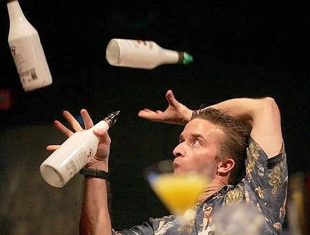 A man at a party juggling bottles with a cocktail in front of him.