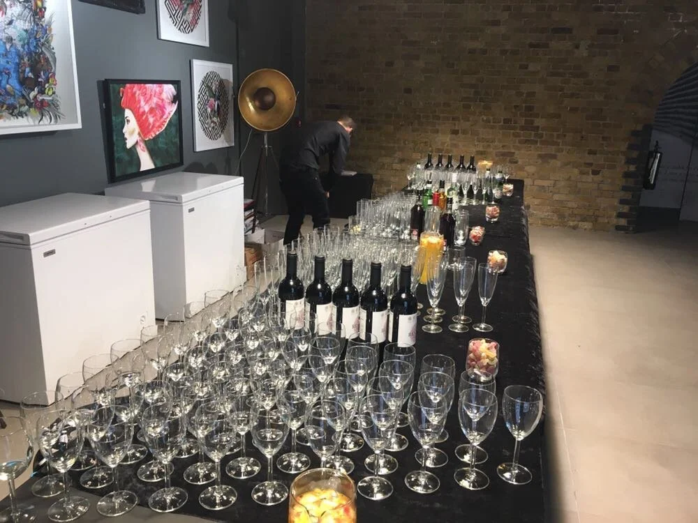 A long table with champagne glasses, wine bottles, and cocktail glasses in a modern art gallery with artwork on the wall and a brick wall in the background.