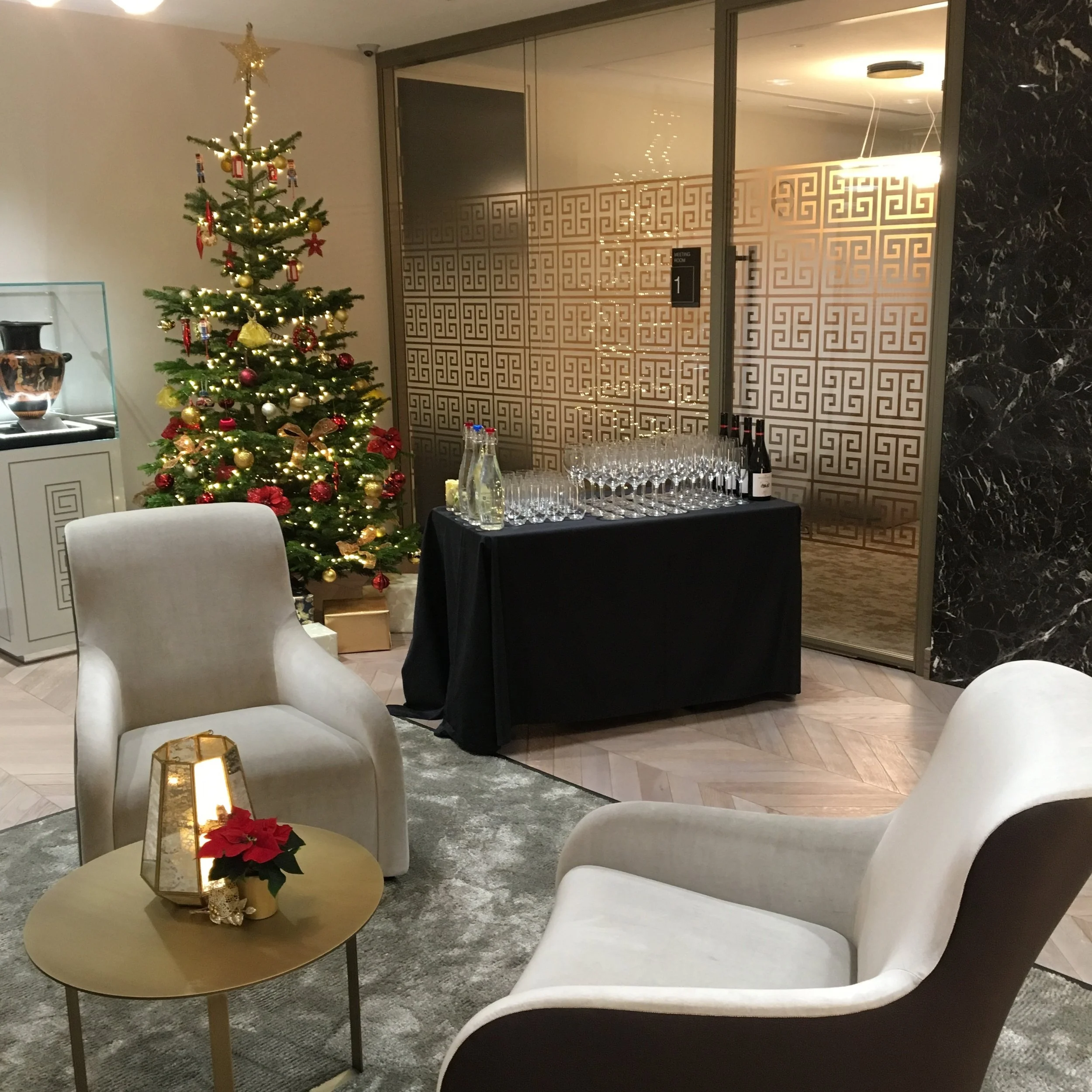 A Christmas tree decorated with red and gold ornaments, topped with a gold star, standing next to a table with glasses and bottles. Two white armchairs and a small gold side table with a poinsettia plant are in a room with modern decor and patterned 
