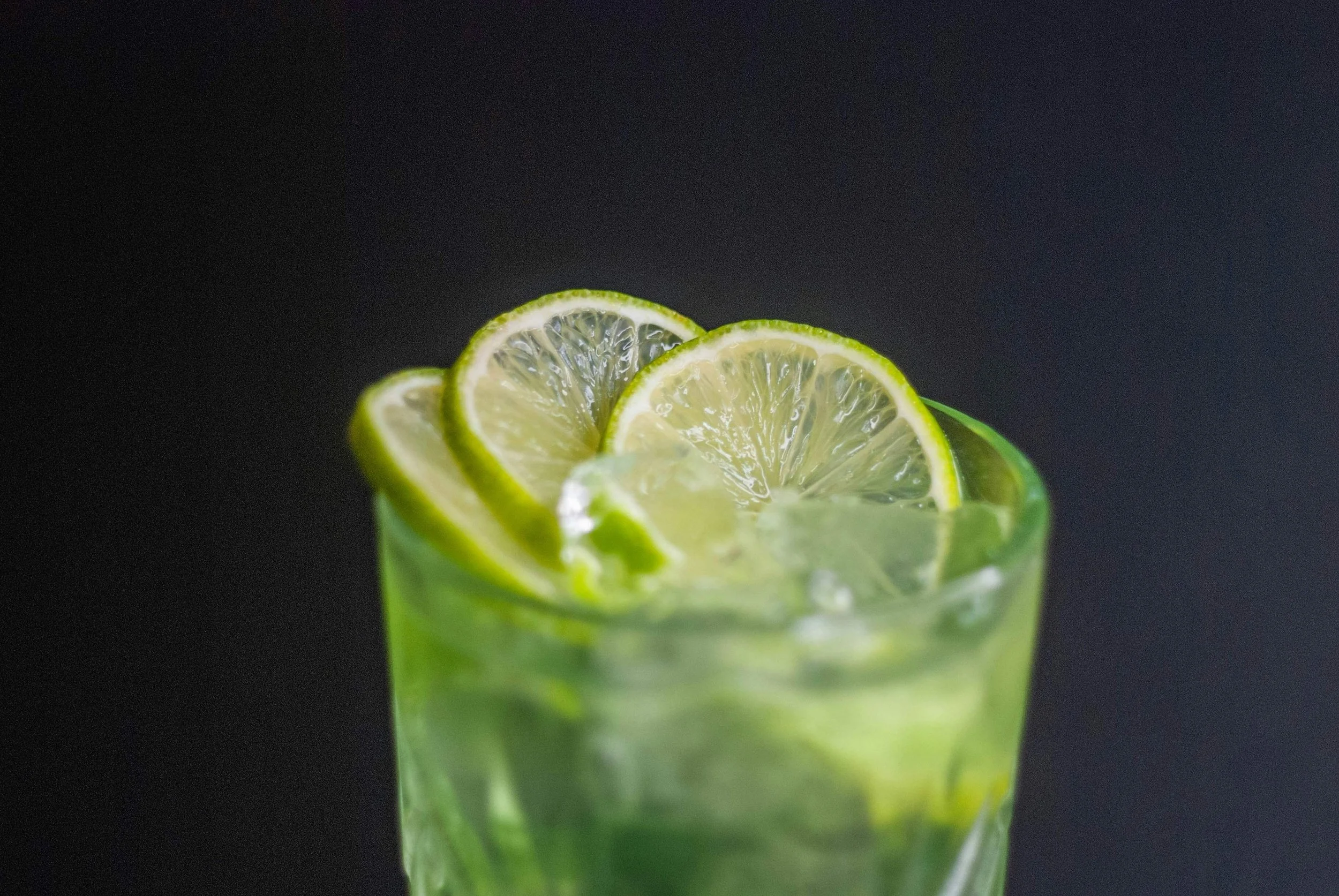 Close-up of a glass with a green drink and lime slices garnish, against a dark background.