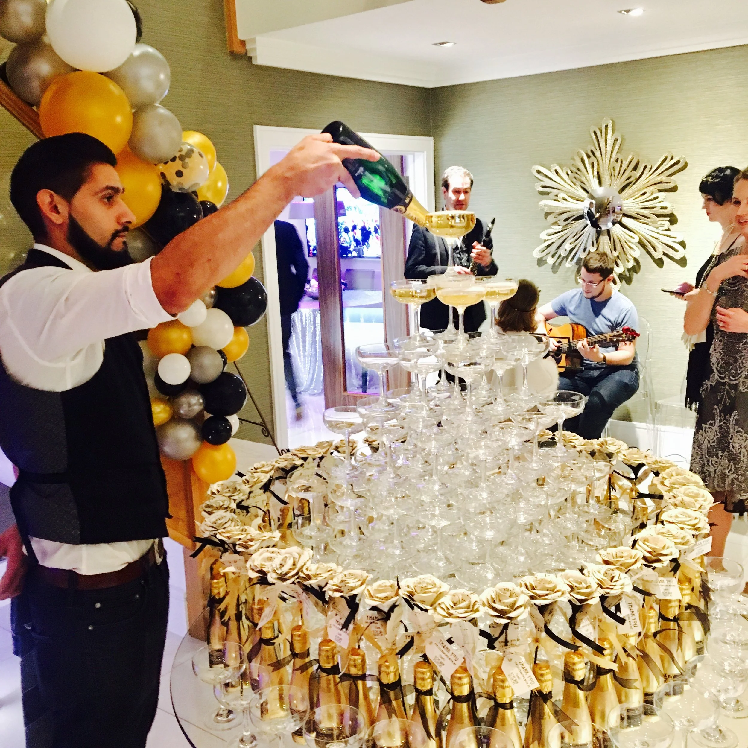 Man pouring champagne into a tower of glasses at a celebration with balloons and musical performance in the background.