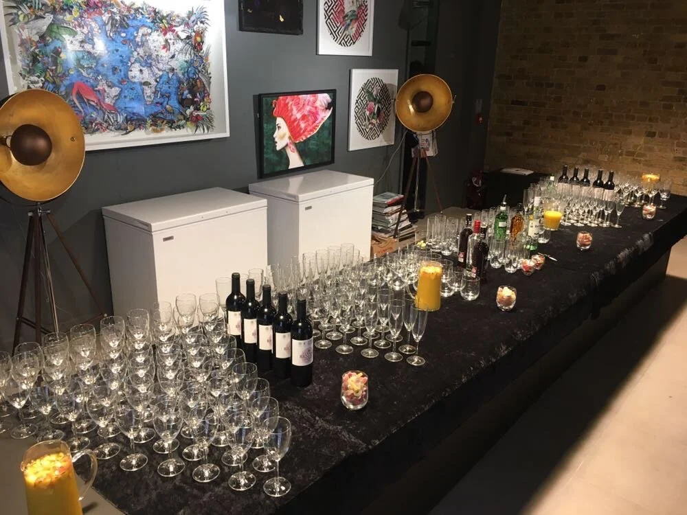 A long table with numerous glasses, bottles, and candles set up for a celebration or event, in a room with artwork and two large floor lamps.
