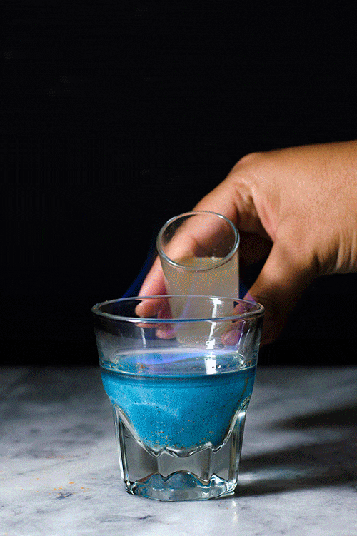 Hand holding a shot glass over a glass of blue liquid on a gray surface with a black background.