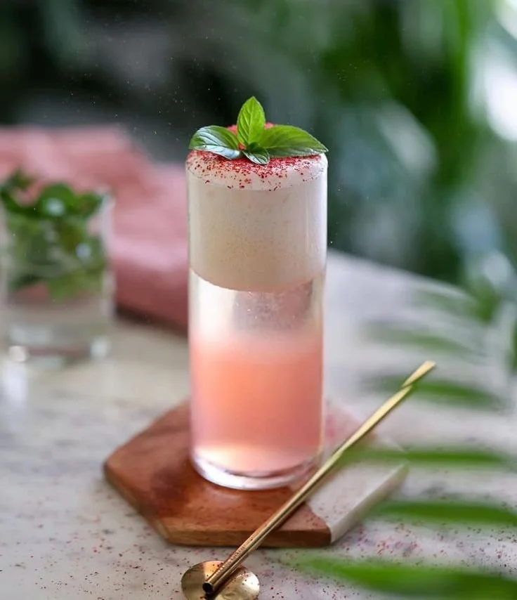 A tall glass with a pink and creamy layered cocktail, garnished with mint leaves and red garnish, on a wooden coaster with a gold straw, on a white surface with green leaves nearby.