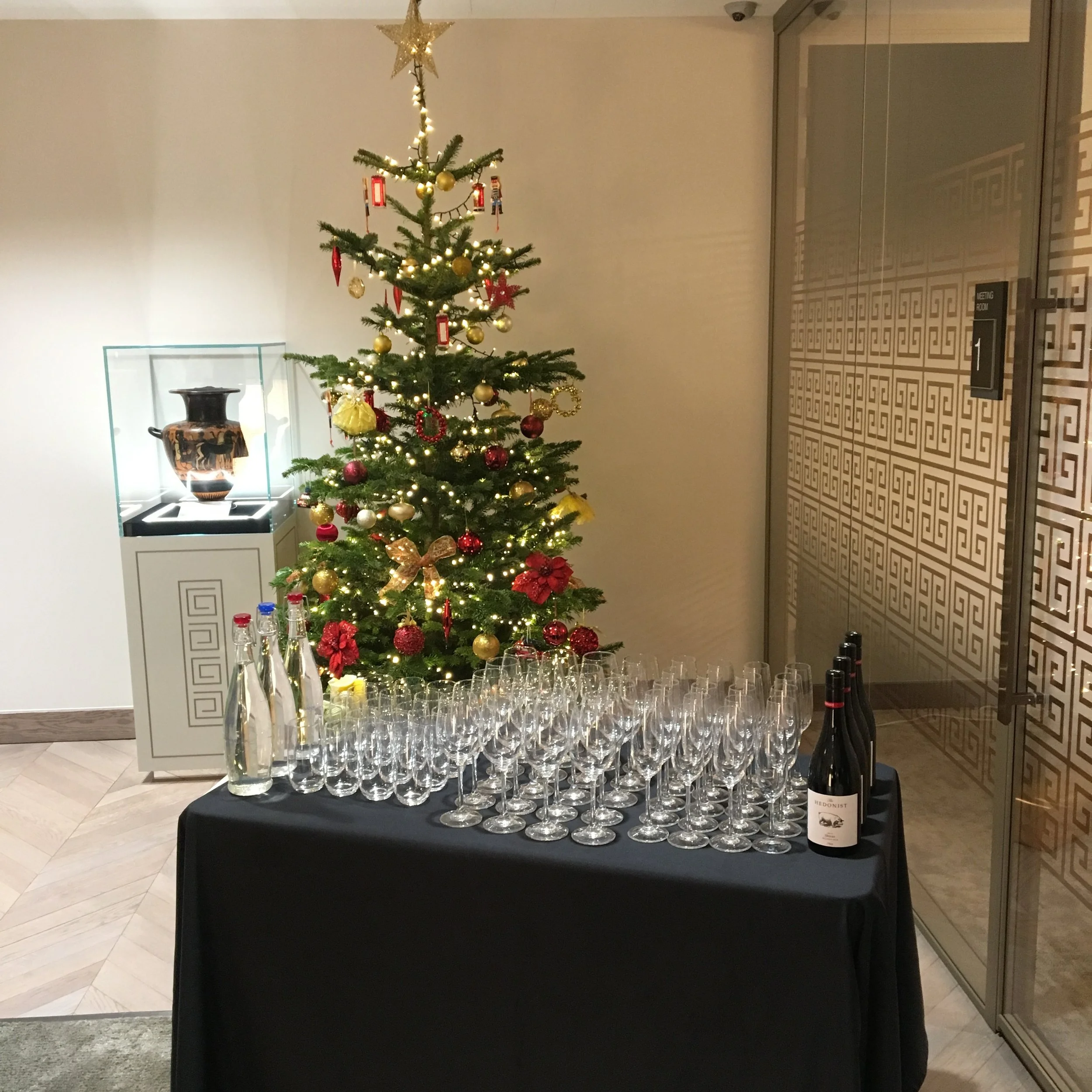 A decorated Christmas tree with gold and red ornaments, lights, and a star on top, next to a table with wine bottles and glasses, in a well-lit room with decorative wall panels.