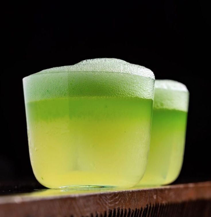 Close-up of two green layered shot glasses with a foamy top, placed on a wooden surface against a black background.