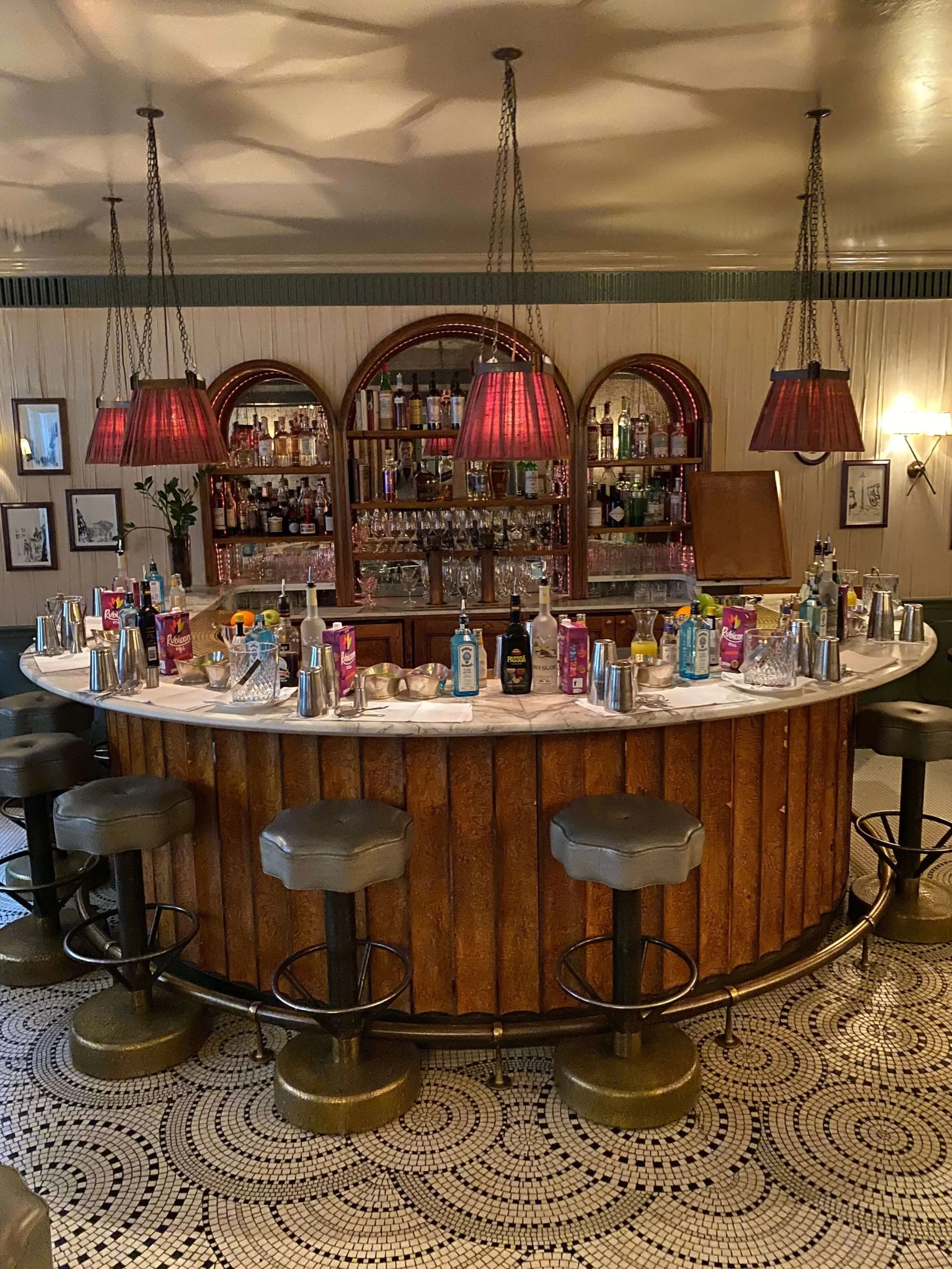 A vintage-style bar with a curved wooden counter, surrounded by four adjustable stools, and a mirrored shelf behind stocked with various liquor bottles and glasses. Hanging red lamps illuminate the bar, with framed pictures on the wall and patterned mosaic tile flooring.