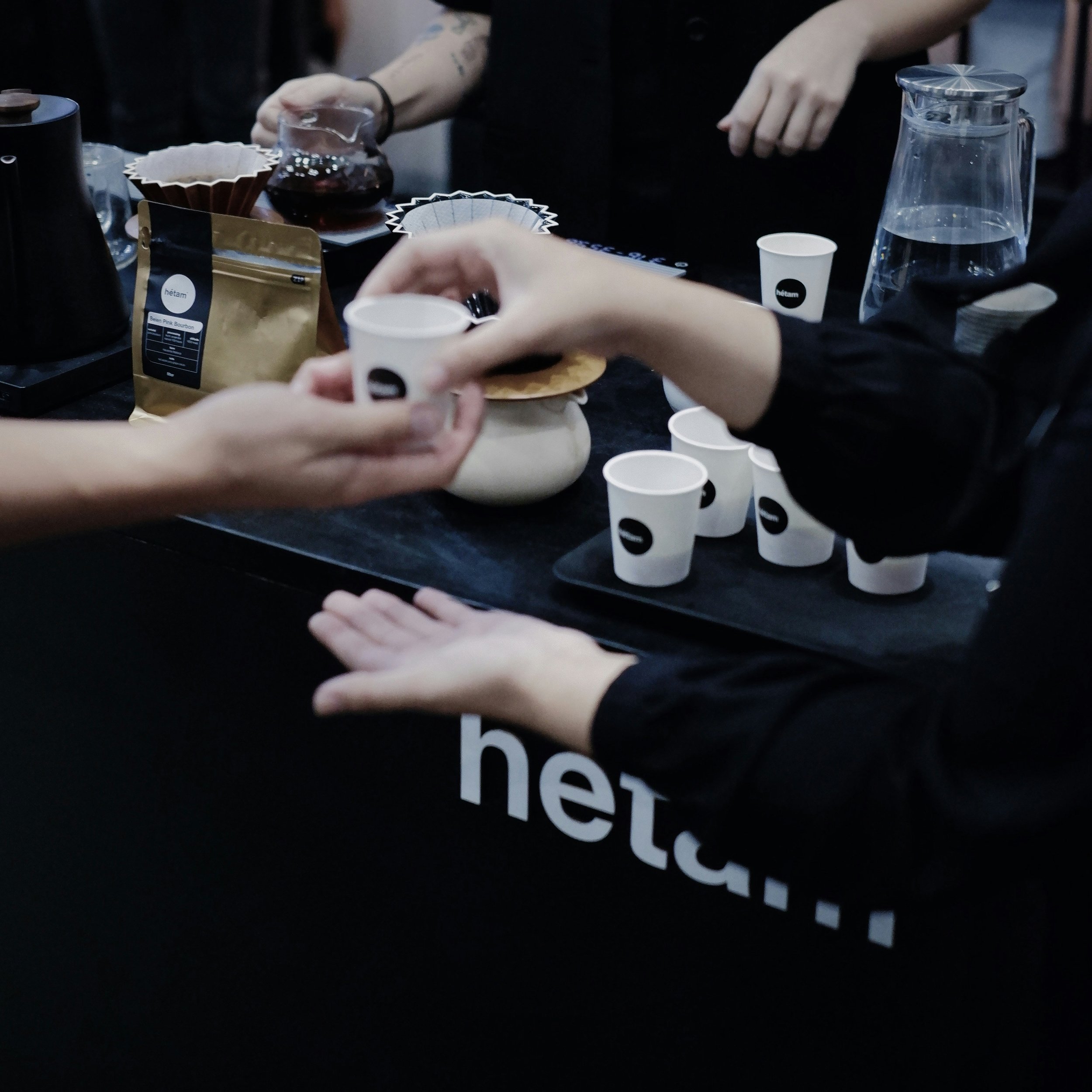People serving and receiving coffee in a coffee shop or tasting event, with cups, a pitcher, and coffee brewing equipment visible.