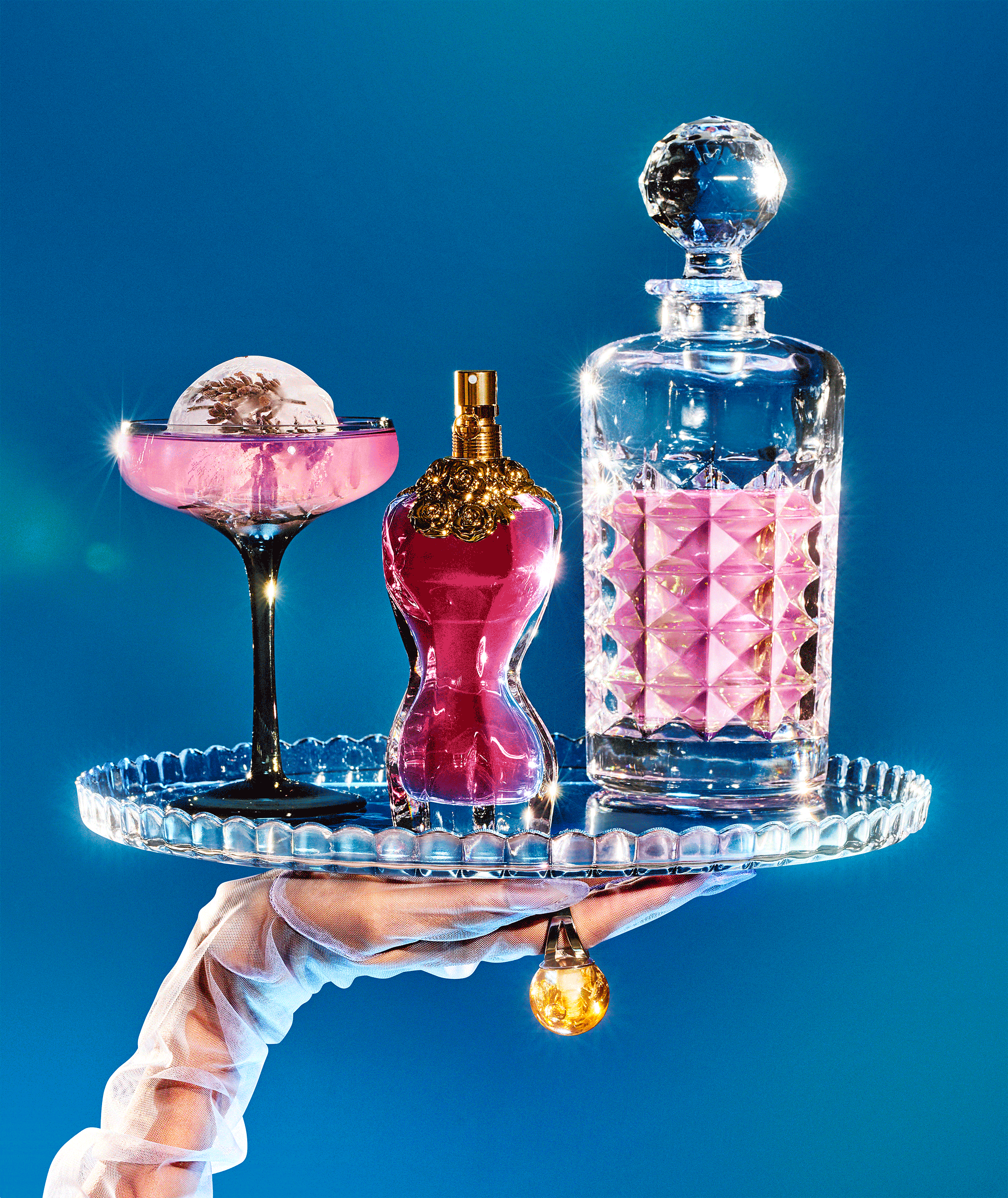 Hand holding a glass tray with three perfume bottles of different shapes and sizes against a blue background.