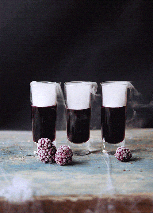Three shot glasses filled with a dark red beverage, with pink berries in front on a rustic wooden surface against a dark background.