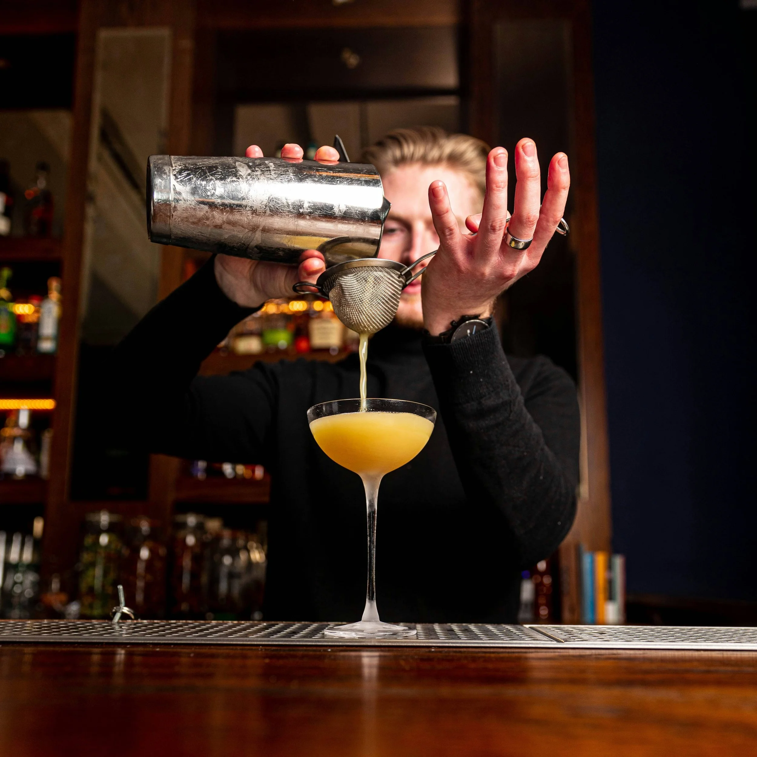 Bartender pouring yellow cocktail through a strainer into a coupe glass at a bar.