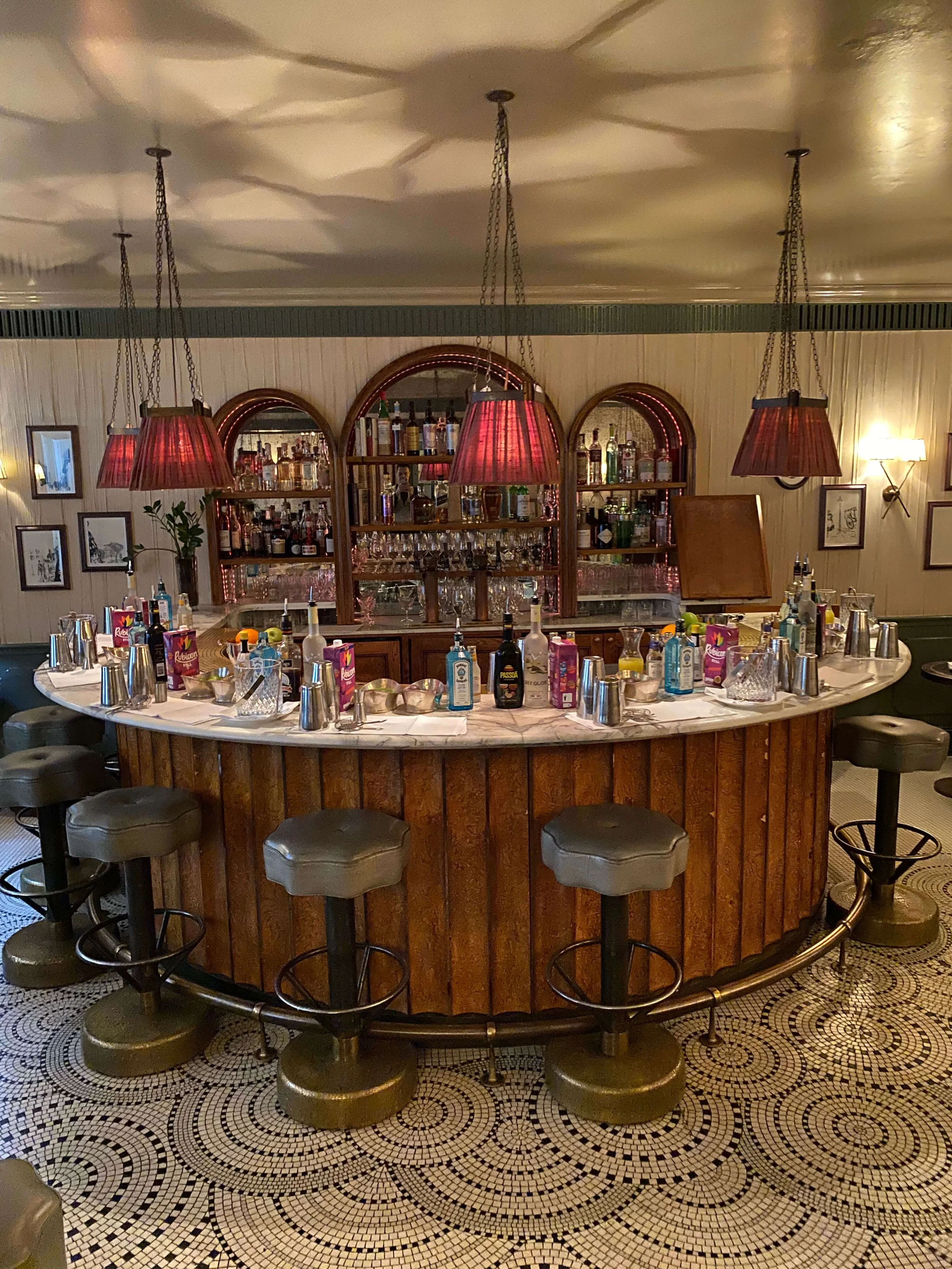 A vintage-style bar with a curved wooden counter, several gray bar stools, and a marble top. The back features wooden shelves with liquor bottles, glasses, and bar tools. Red pendant lamps hang from the ceiling, casting shadows on the ceiling. The fl