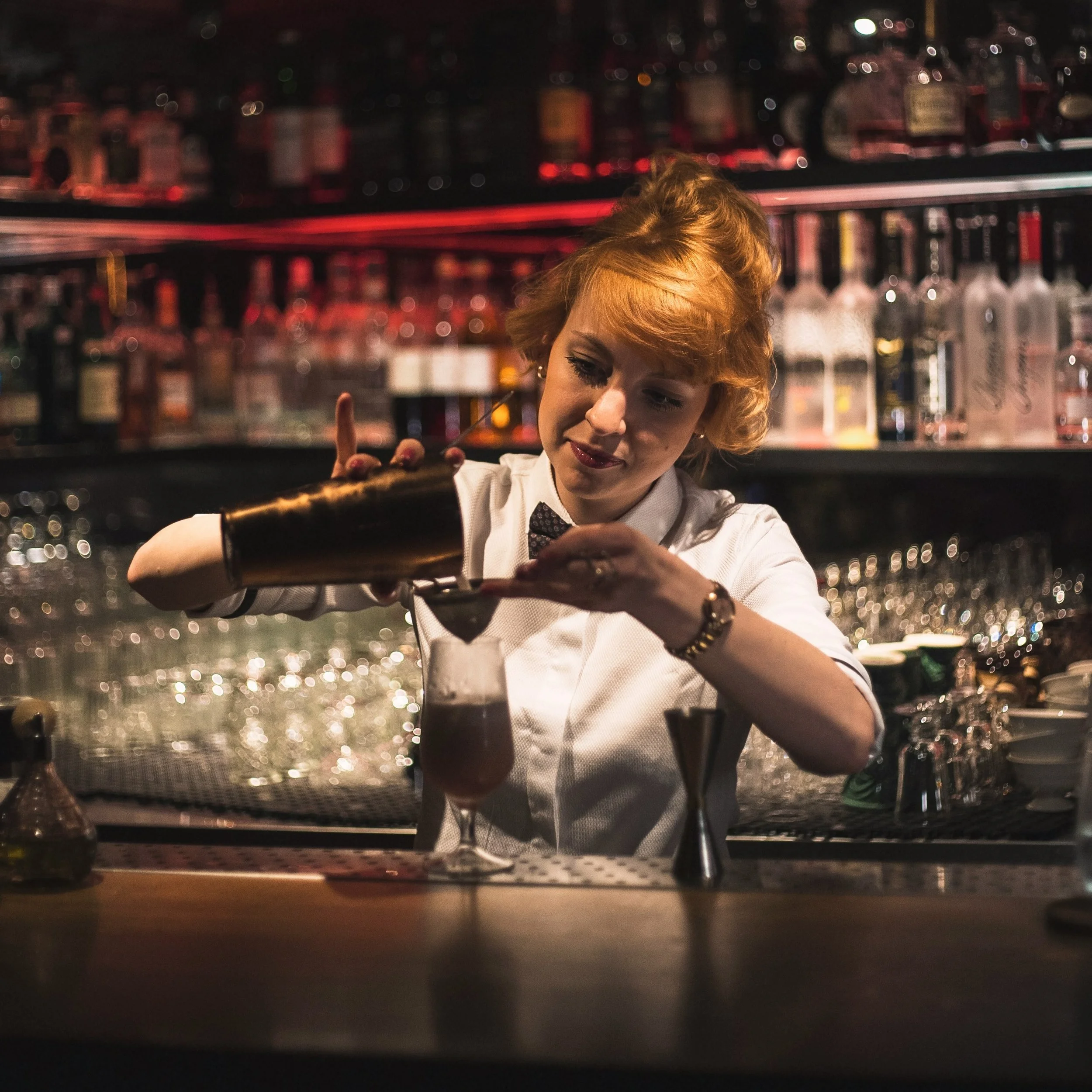 A woman with short curly red hair and a white shirt is in a bar, looking at her phone. The background shows shelves of liquor bottles illuminated with red and warm lighting.