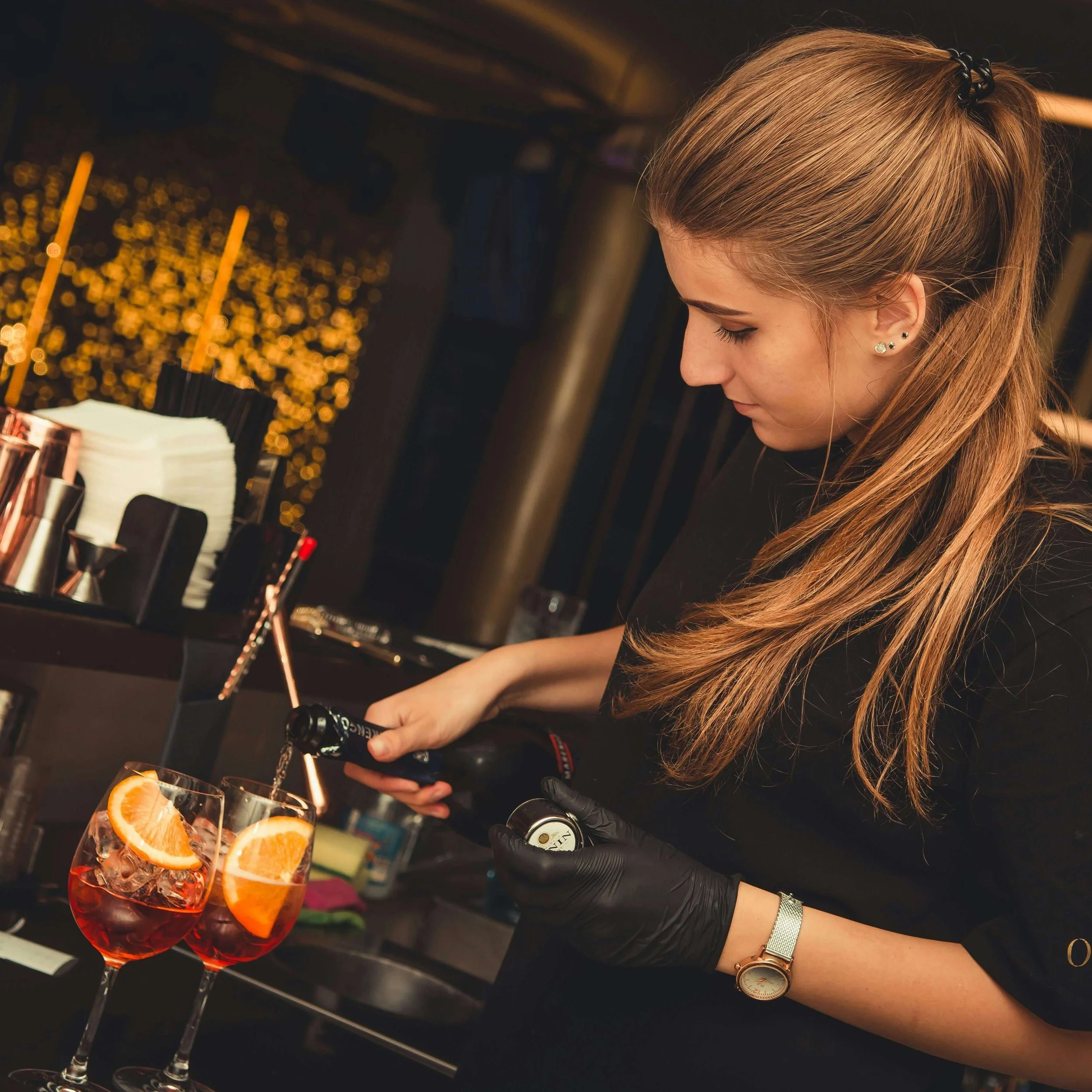 A woman with long, light brown hair tied back, wearing a black shirt, a watch, and black gloves, is pouring a dark liquor from a bottle into a glass with ice and orange slices at a bar.