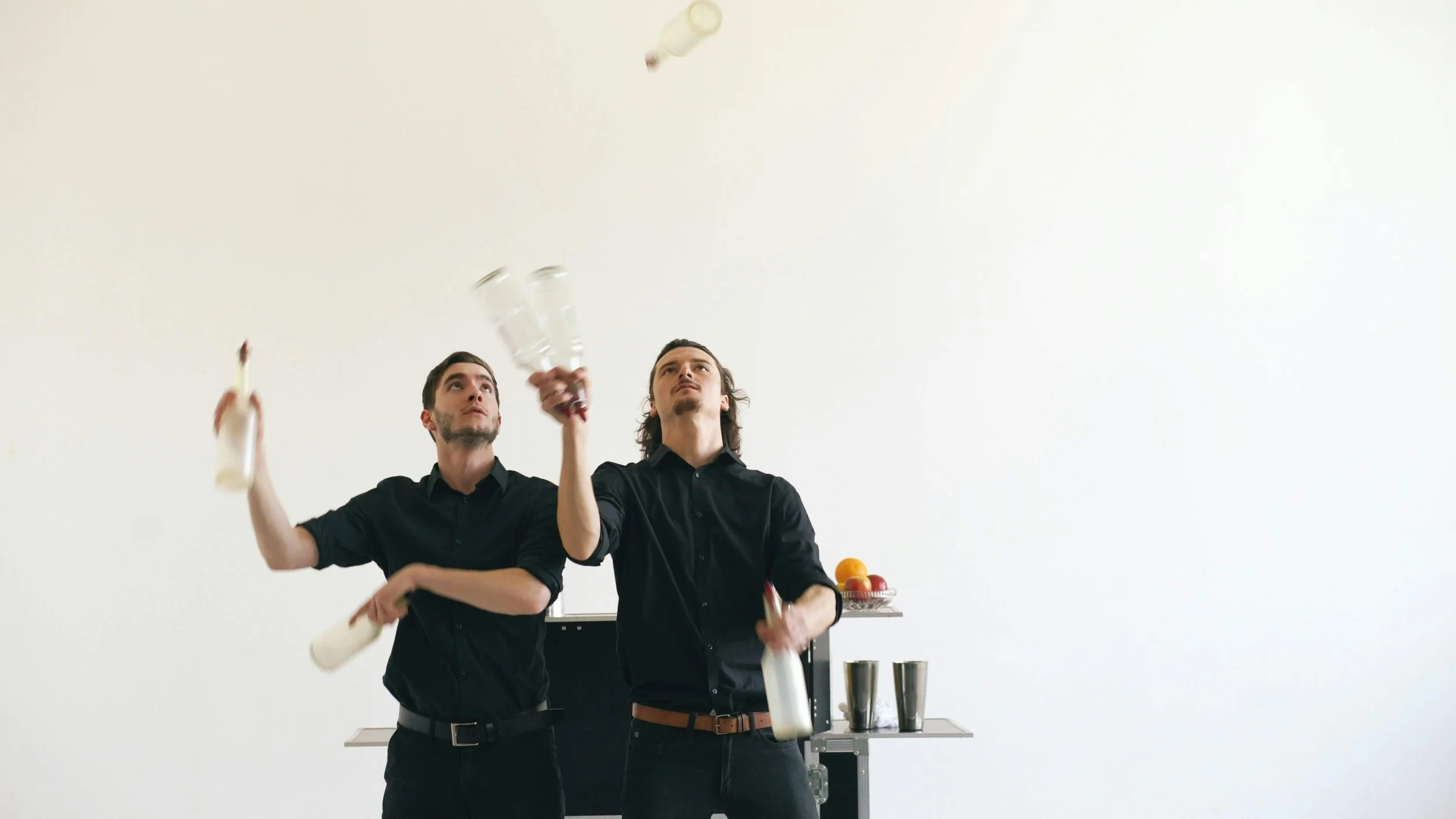 Two men in black shirts juggling bottles and glasses in a white room with a fruit basket and metal cups in the background.